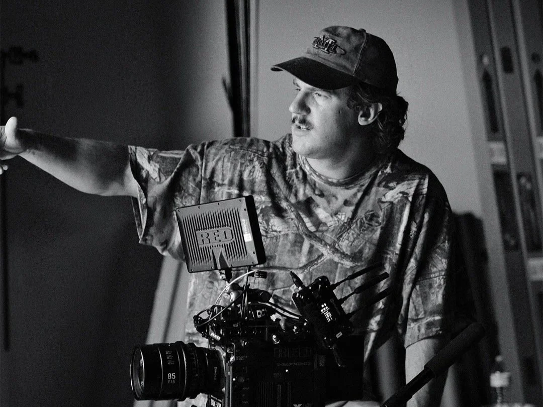 A man dressed in camouflage shirt and wearing a cap, operating a professional video camera setup, pointing to the side and looking in that direction, in an indoor setting.