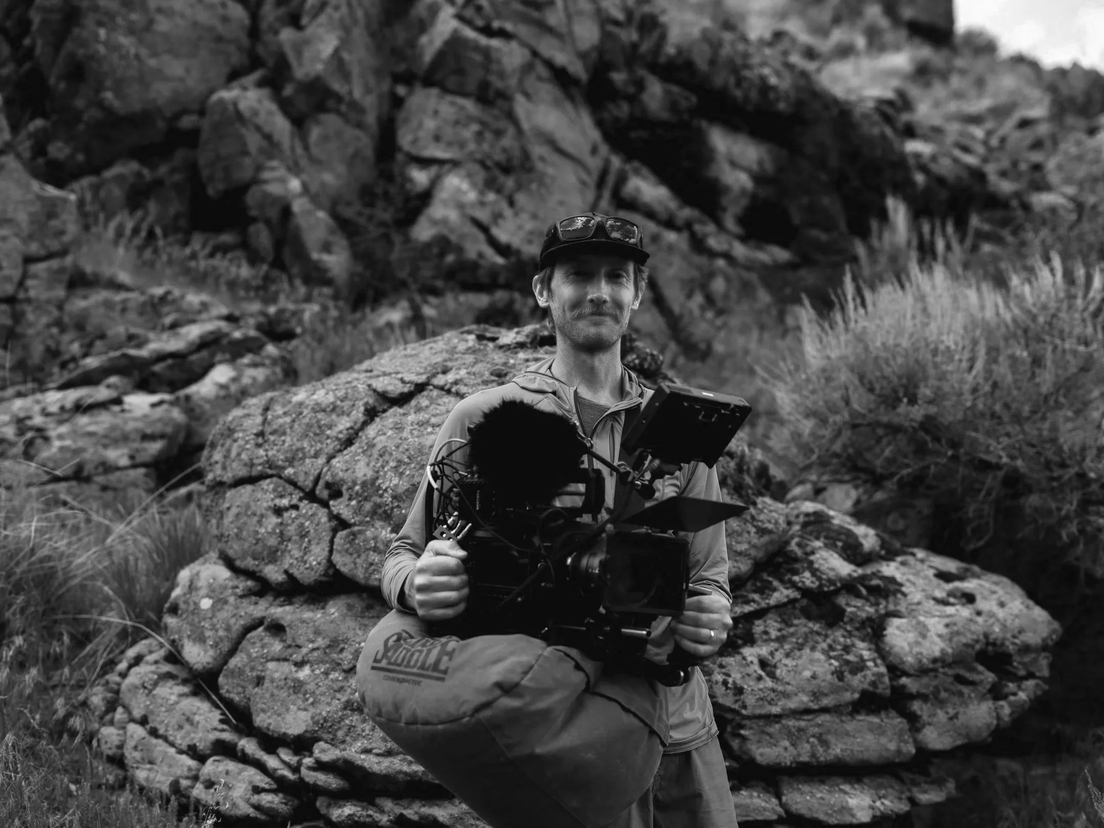 A man in outdoor gear holds a professional camera on a tripod, standing outdoors with a cityscape in the background.