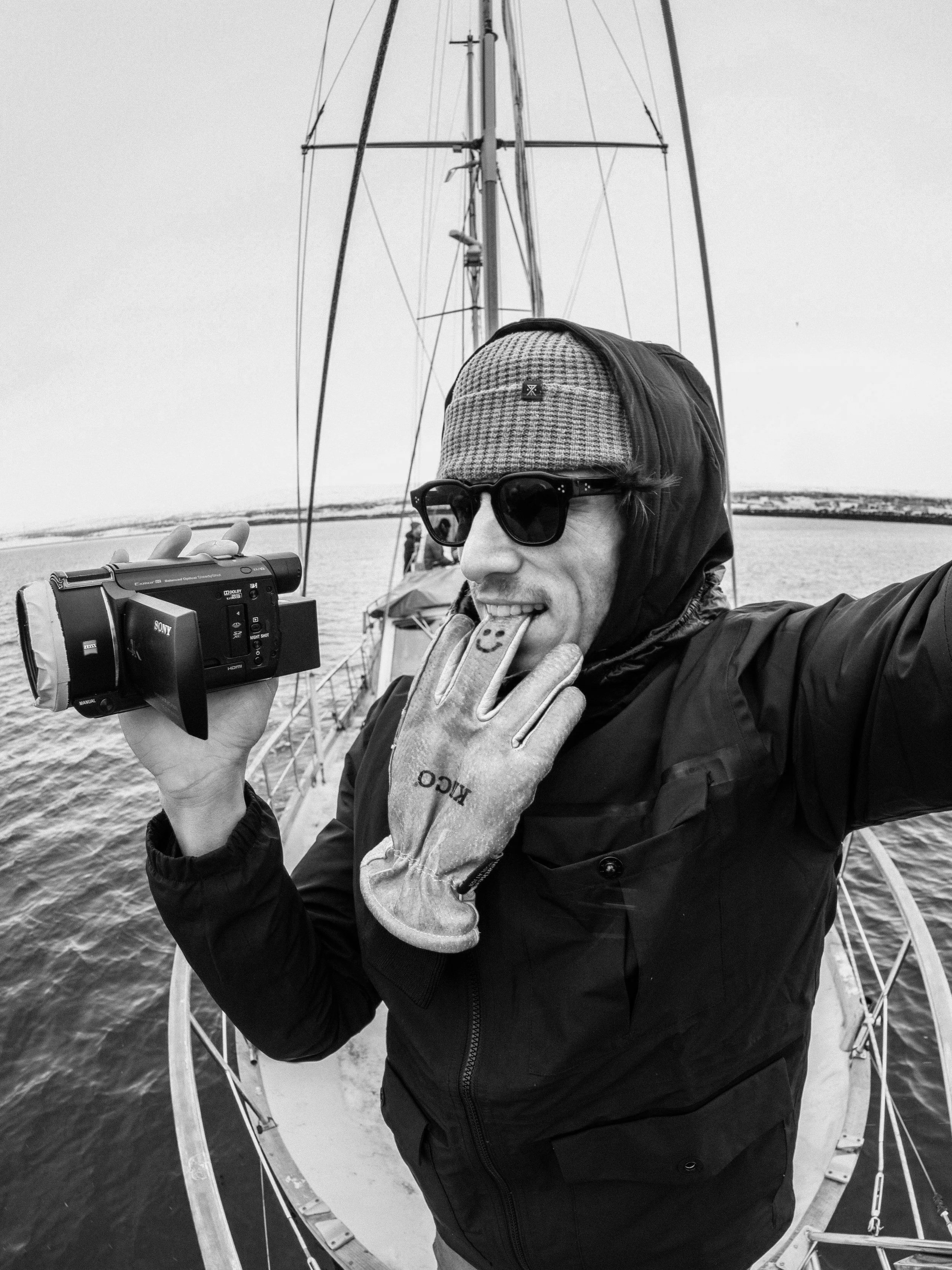 Person on a boat holding a camera and making a playful face, wearing sunglasses, gloves, and a jacket, with water and boat rigging in the background.