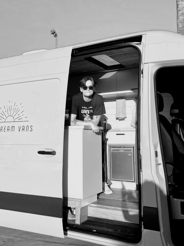 A person with sunglasses, wearing a graphic t-shirt, leaning on the counter inside a small camper van or food truck, with the side door open showing the interior kitchenette, outdoors on a sunny day.