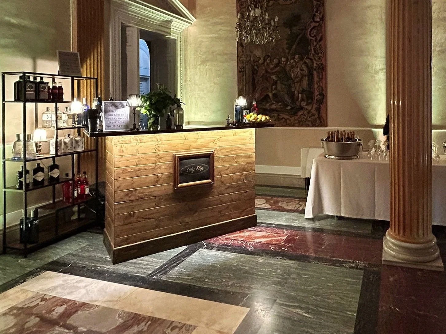 Un'area bar con un bancone di legno e scaffali con bottiglie di bevande. Sul bancone ci sono piante e lampade. Sullo sfondo, un quadro e un tavolo con una ciotola di bottiglie, bicchieri e altri oggetti.