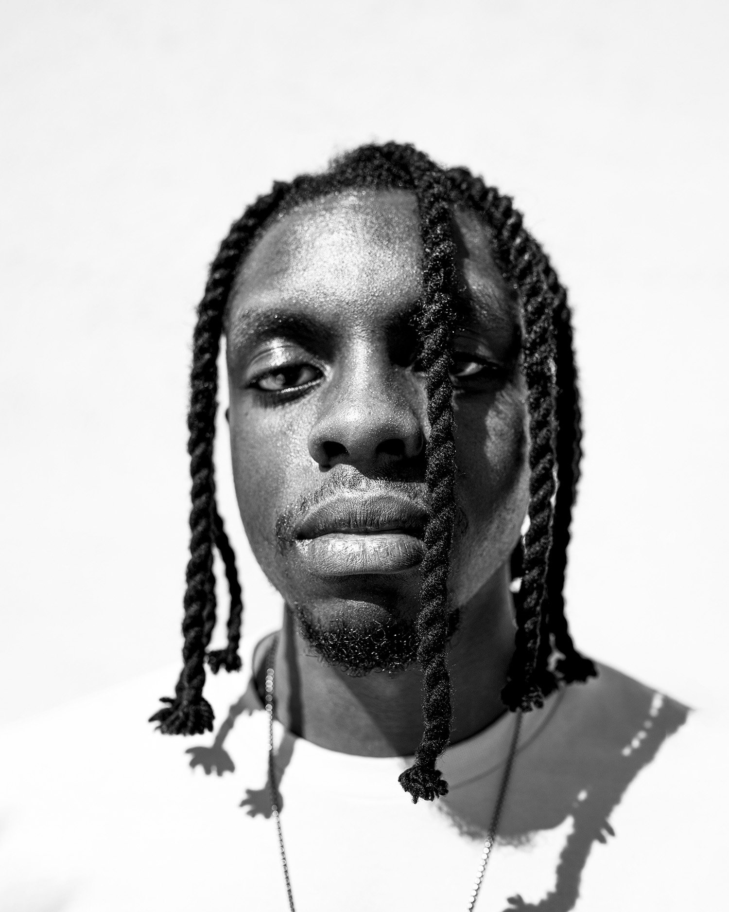 Close-up black and white portrait of a man with braided hair, looking directly at the camera, against a plain background.
