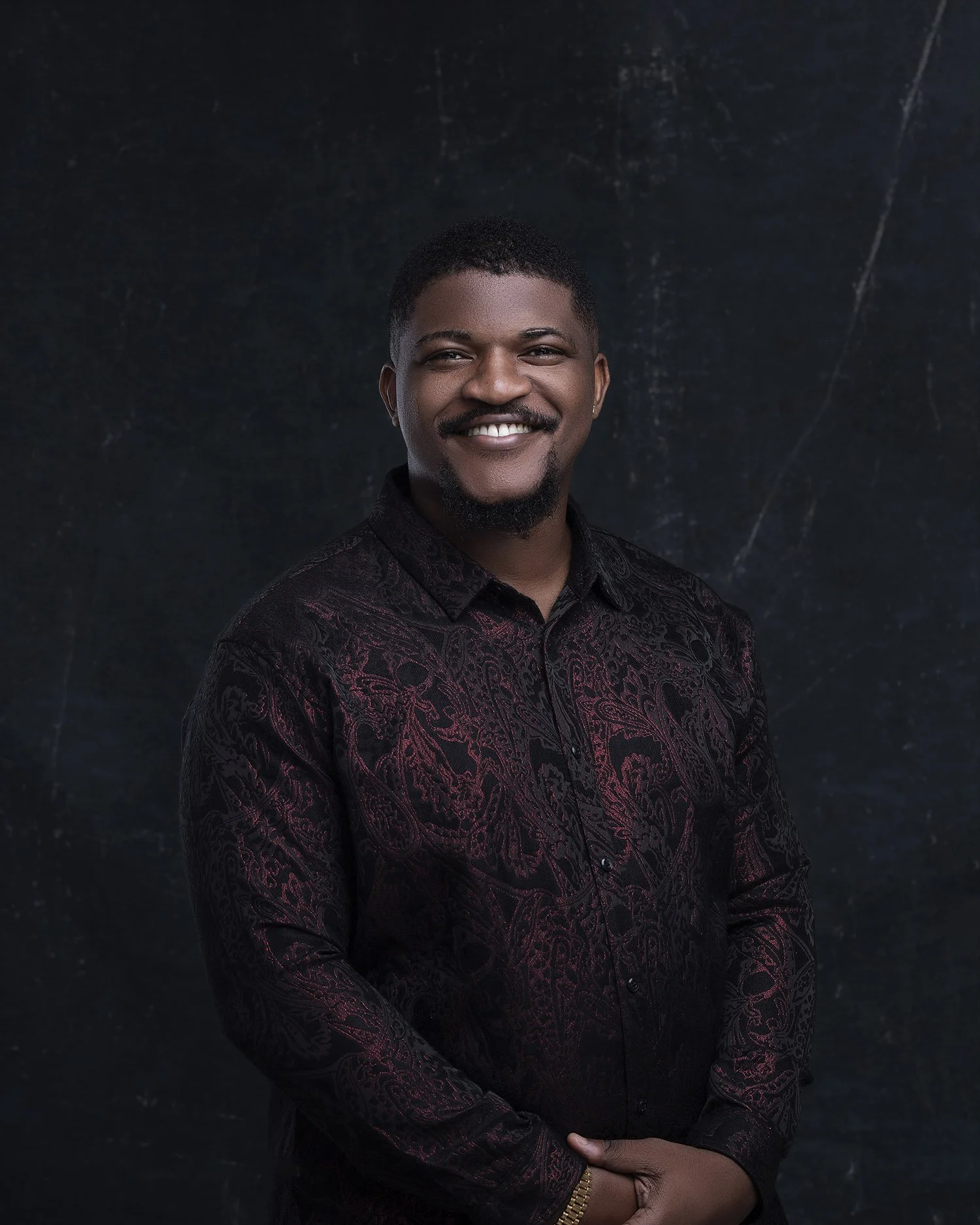 A young man with a short haircut and goatee smiling, dressed in a dark patterned shirt, standing against a dark background.