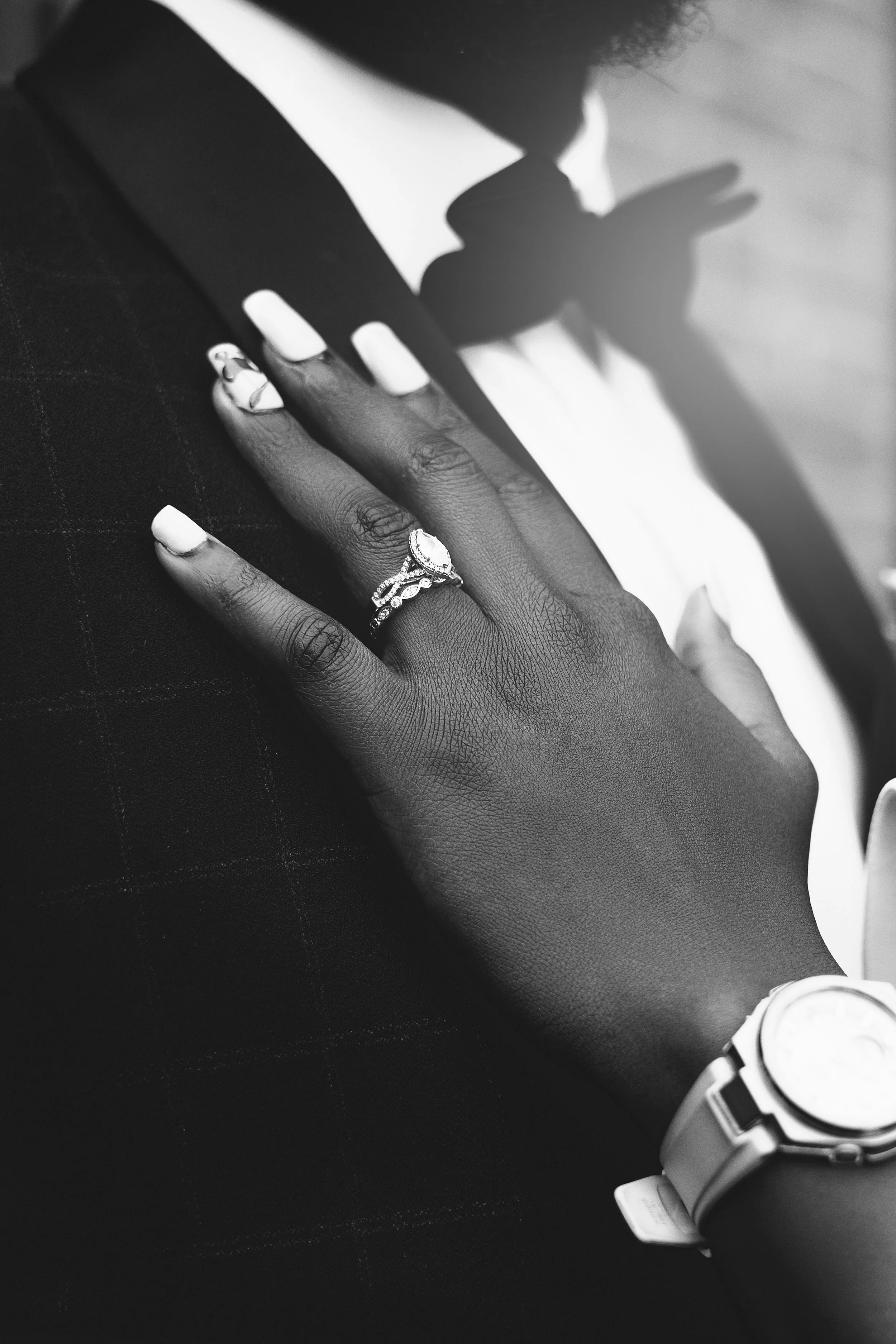 Close-up of a person with dark skin wearing a ring and a watch, touching another person in a tuxedo, in black and white.