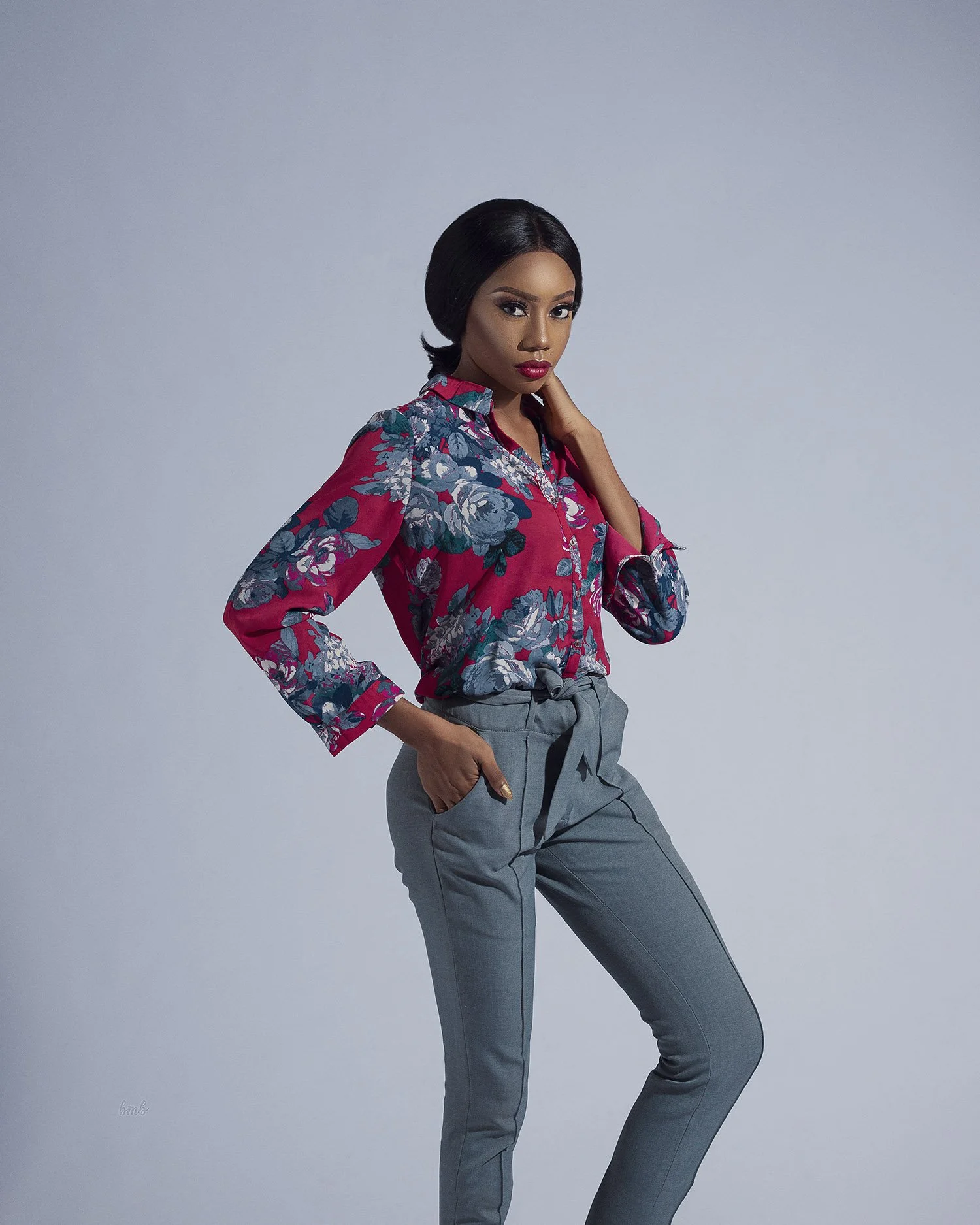 A woman with dark hair and makeup, wearing a red floral blouse and gray high-waisted pants, posing against a light gray background.