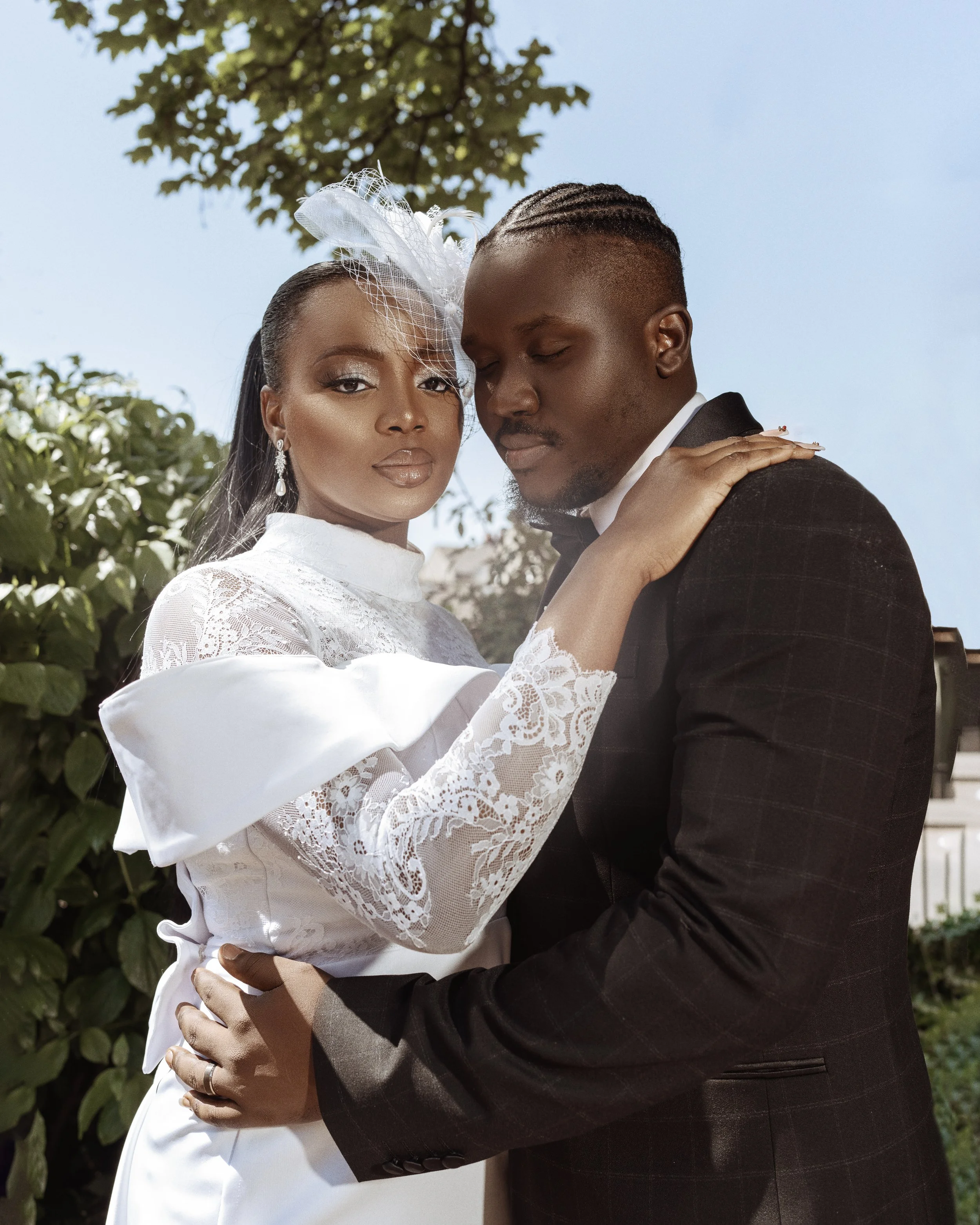 A bride and groom embrace outdoors, with the bride dressed in a white lace dress with puffed sleeves and a fascinator, and the groom in a dark suit with a white shirt, against a backdrop of green foliage and a blue sky.