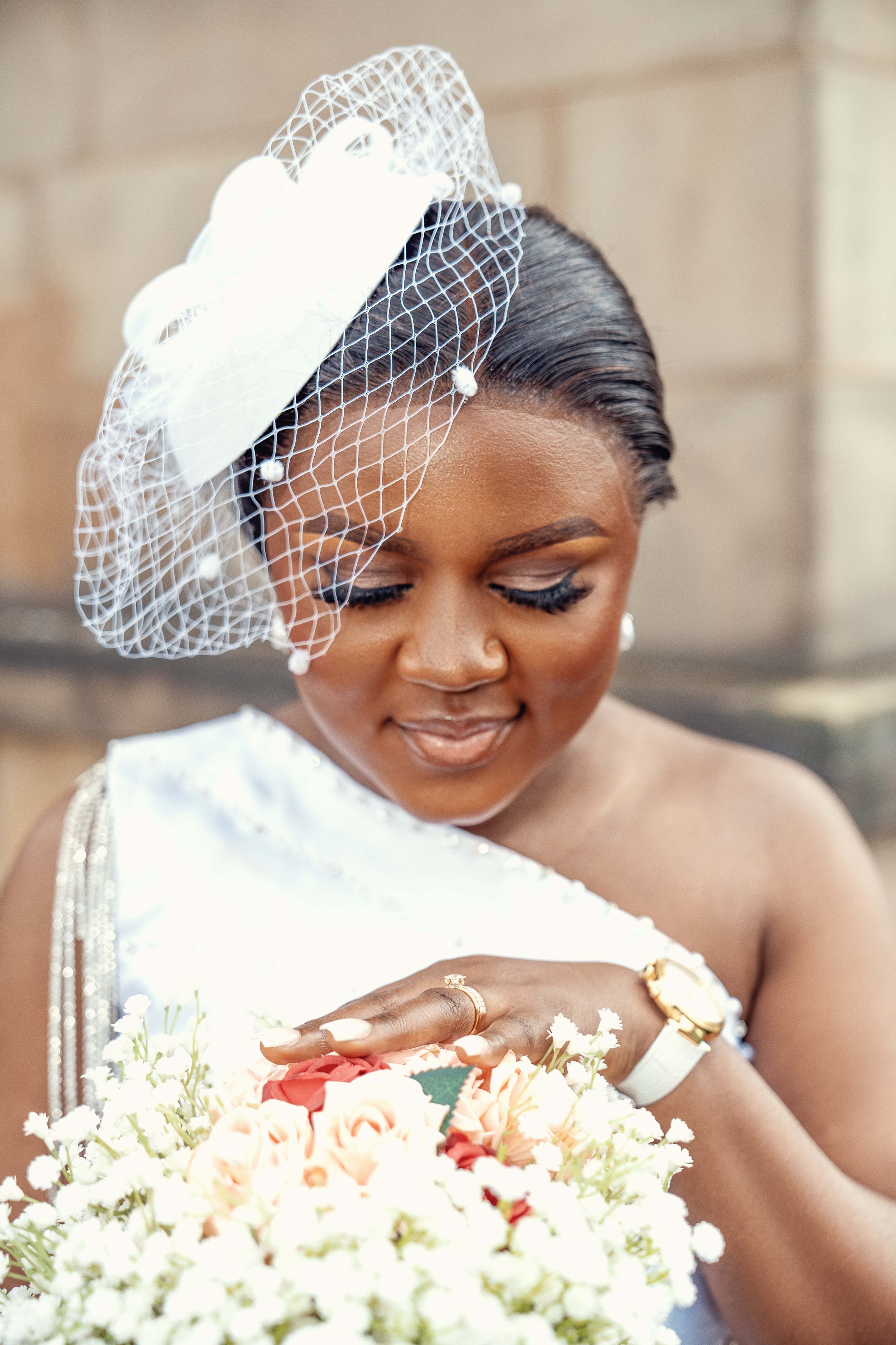 A woman in a white dress with a fascinator hat holding a bouquet of flowers, looking down with a gentle smile.