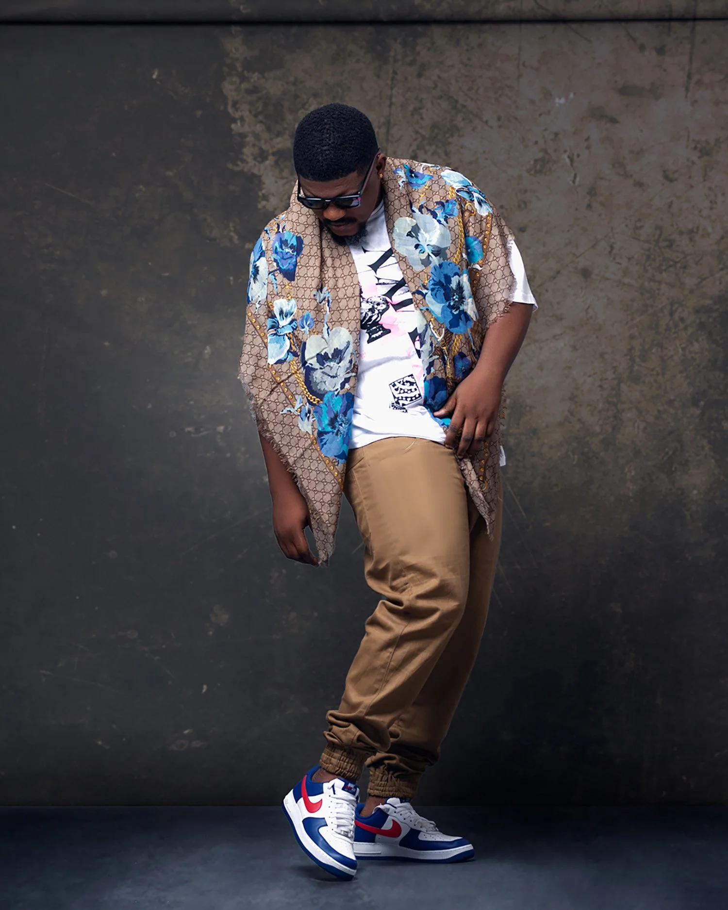 A man wearing sunglasses poses against a dark grey background, dressed in a floral-patterned coat, graphic t-shirt, beige jogger pants, and Nike sneakers with red, white, and blue accents.