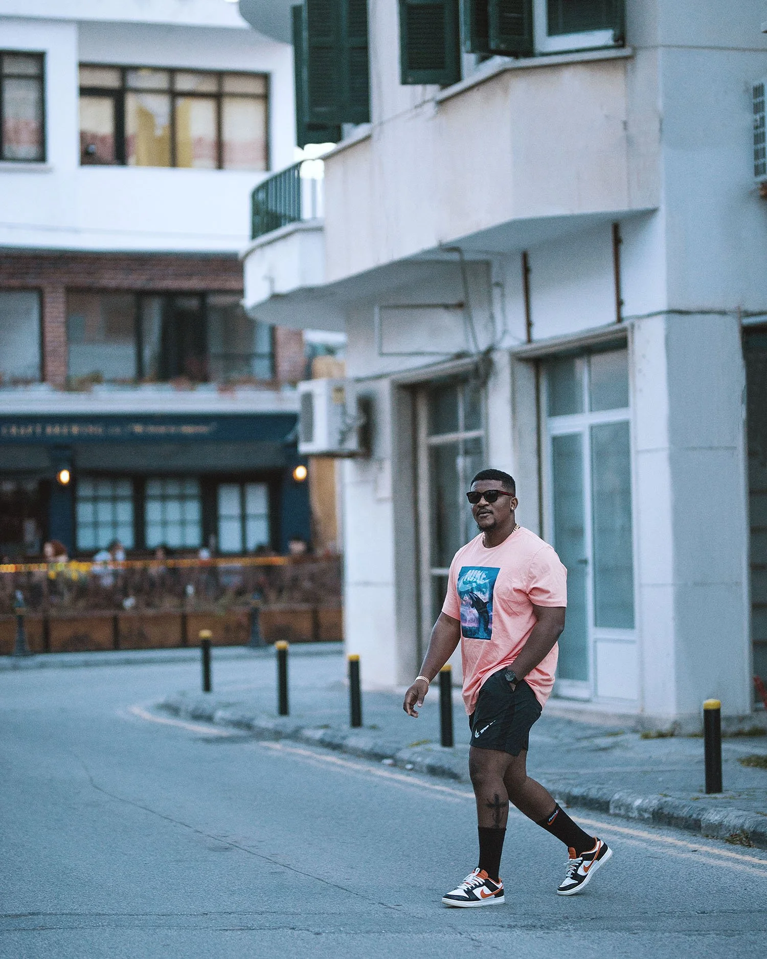 A man walking on a city street wearing a pink Nike t-shirt, black shorts, black socks, and sneakers, with sunglasses and a watch, with buildings in the background.