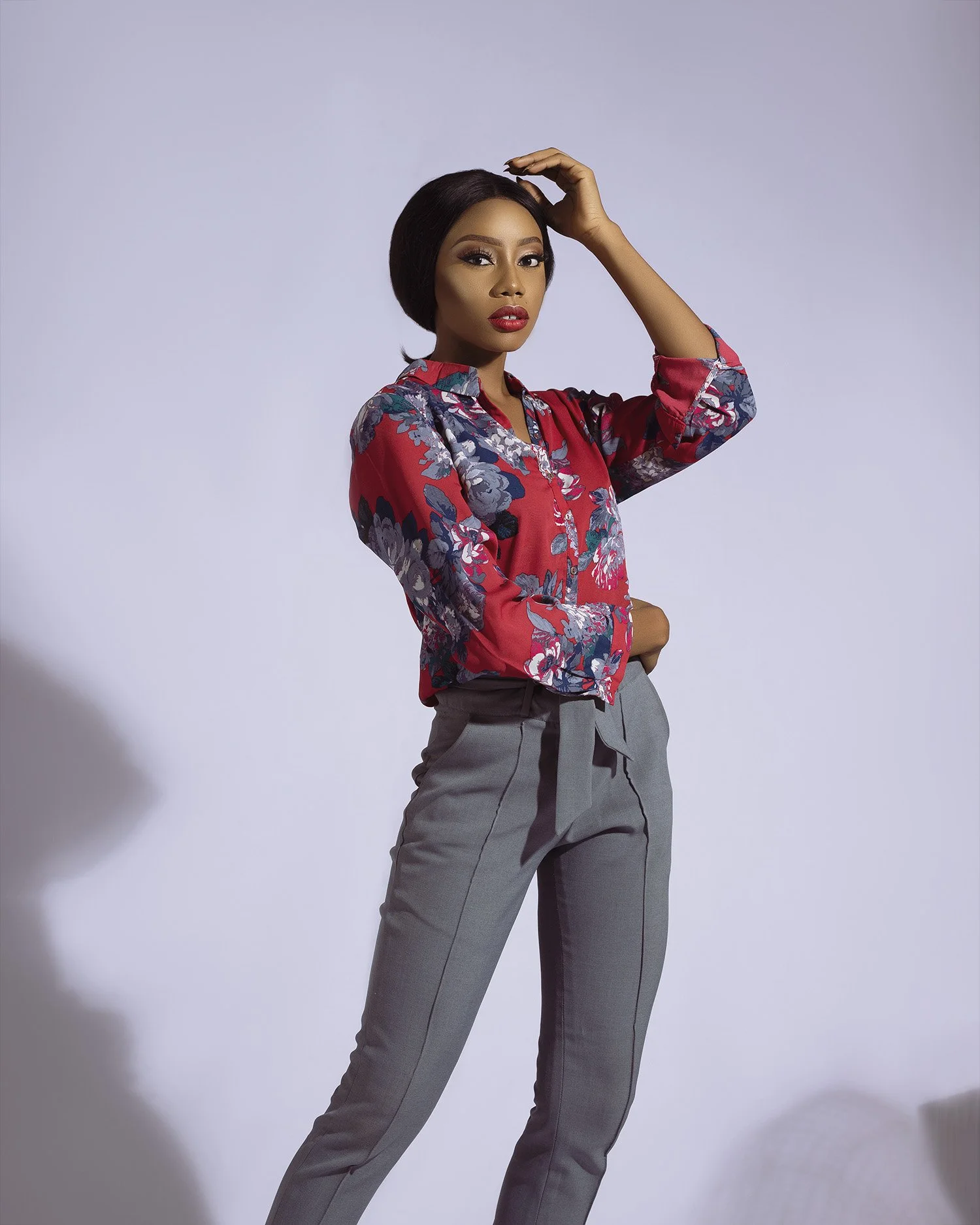 A woman with dark hair styled in a bob, wearing a colorful floral blouse and gray tailored pants, poses against a plain background.