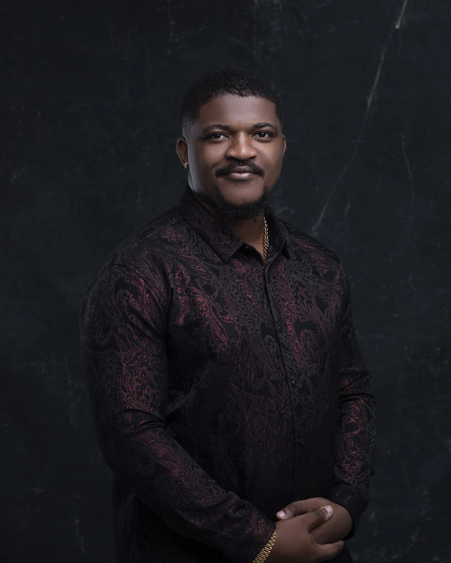A young man with a dark, patterned shirt, gold jewelry, and a slight smile stands against a dark, textured background.