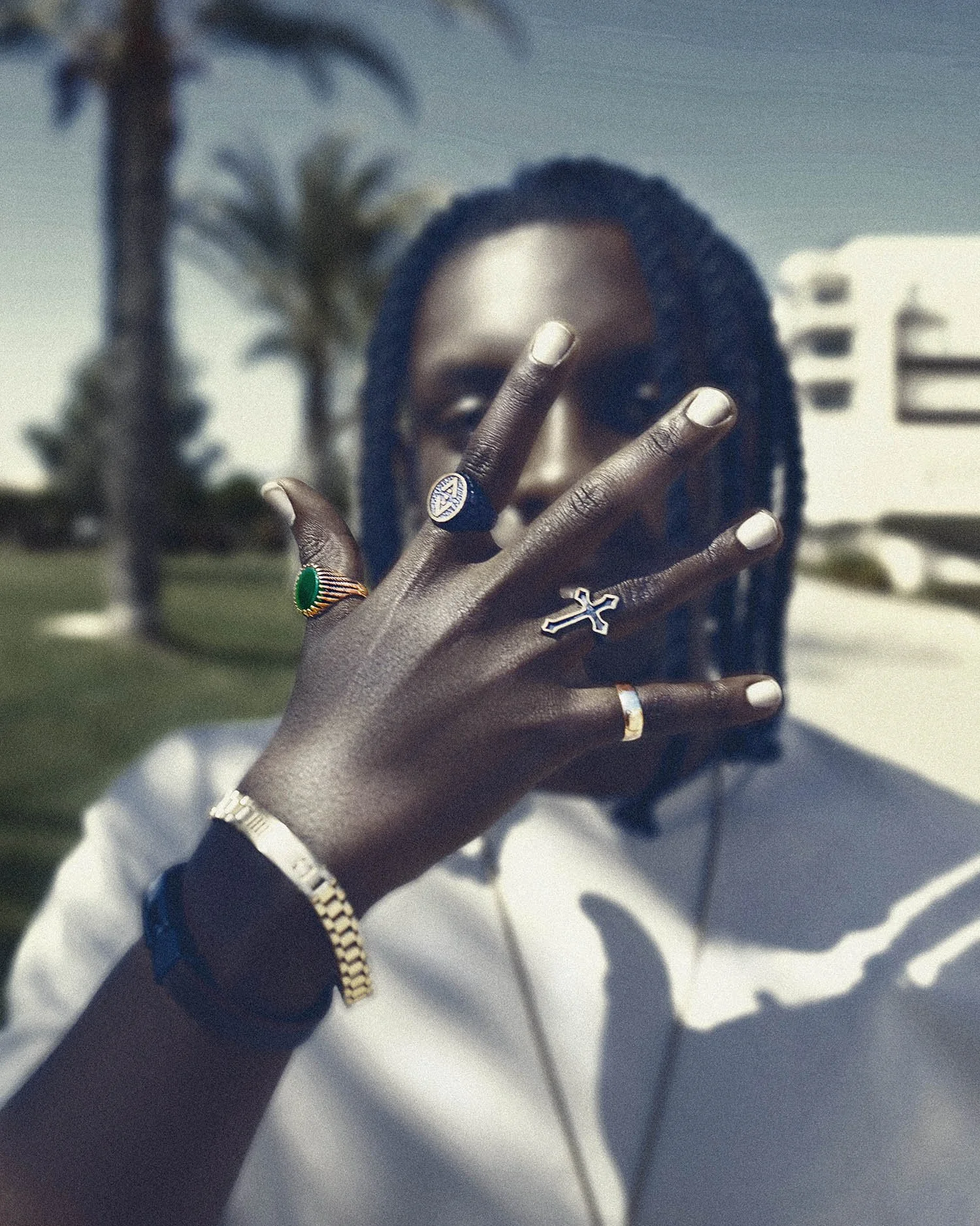 A woman with braided hair holds her hand up, showing rings and a gold watch on her wrist. She is outdoors with palm trees and modern white buildings in the background.