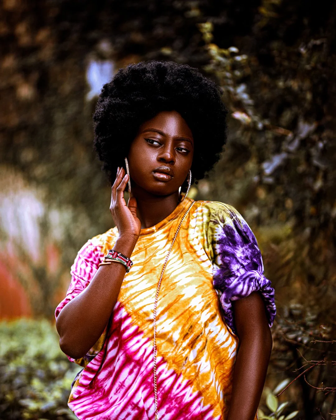 A woman with dark skin and natural curly hair standing outdoors in a forested area, wearing a colorful tie-dye shirt in yellow, pink, purple, and white, accessorized with hoop earrings, bracelets, and a necklace.