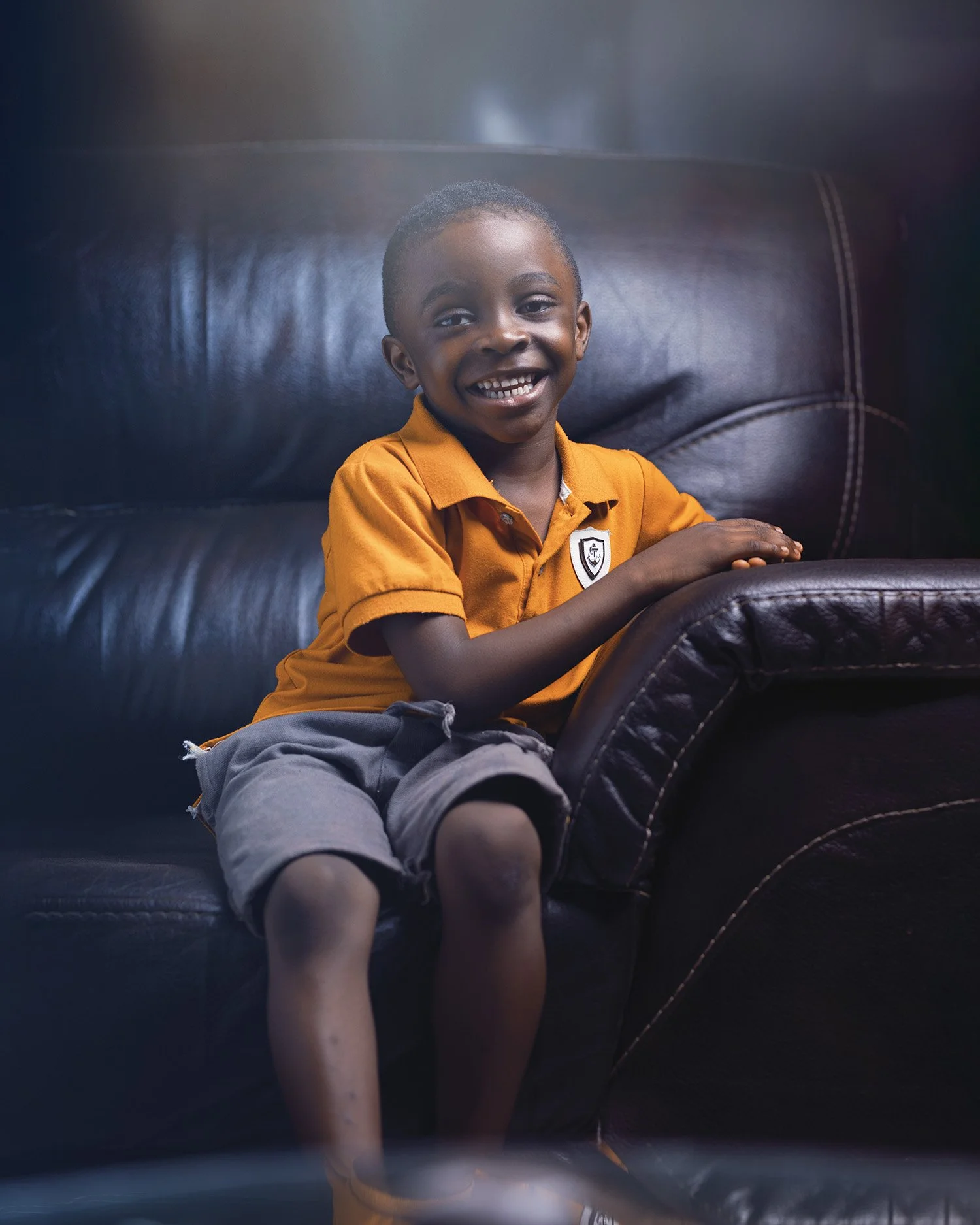 A young boy with a big smile, wearing an orange polo shirt and gray shorts, sitting on a dark leather couch.