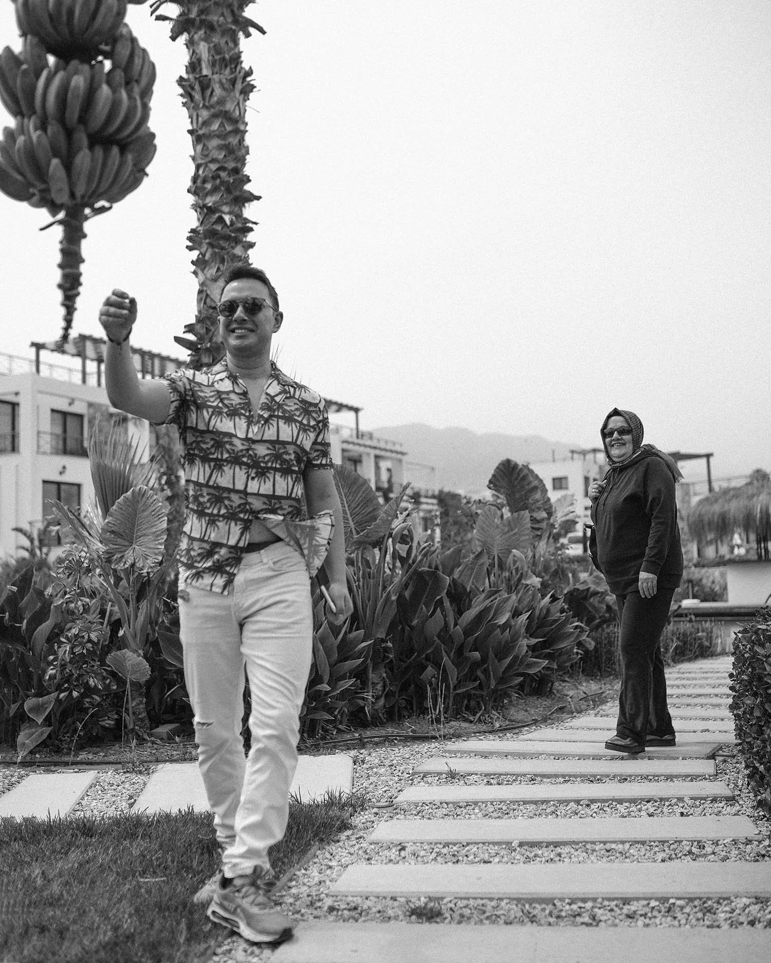 Two people walking outdoors on a path with plants and buildings in the background. The man in front is smiling, wearing glasses, a patterned shirt, and light-colored pants. The woman behind is smiling, wearing sunglasses, a headscarf, and a dark outf