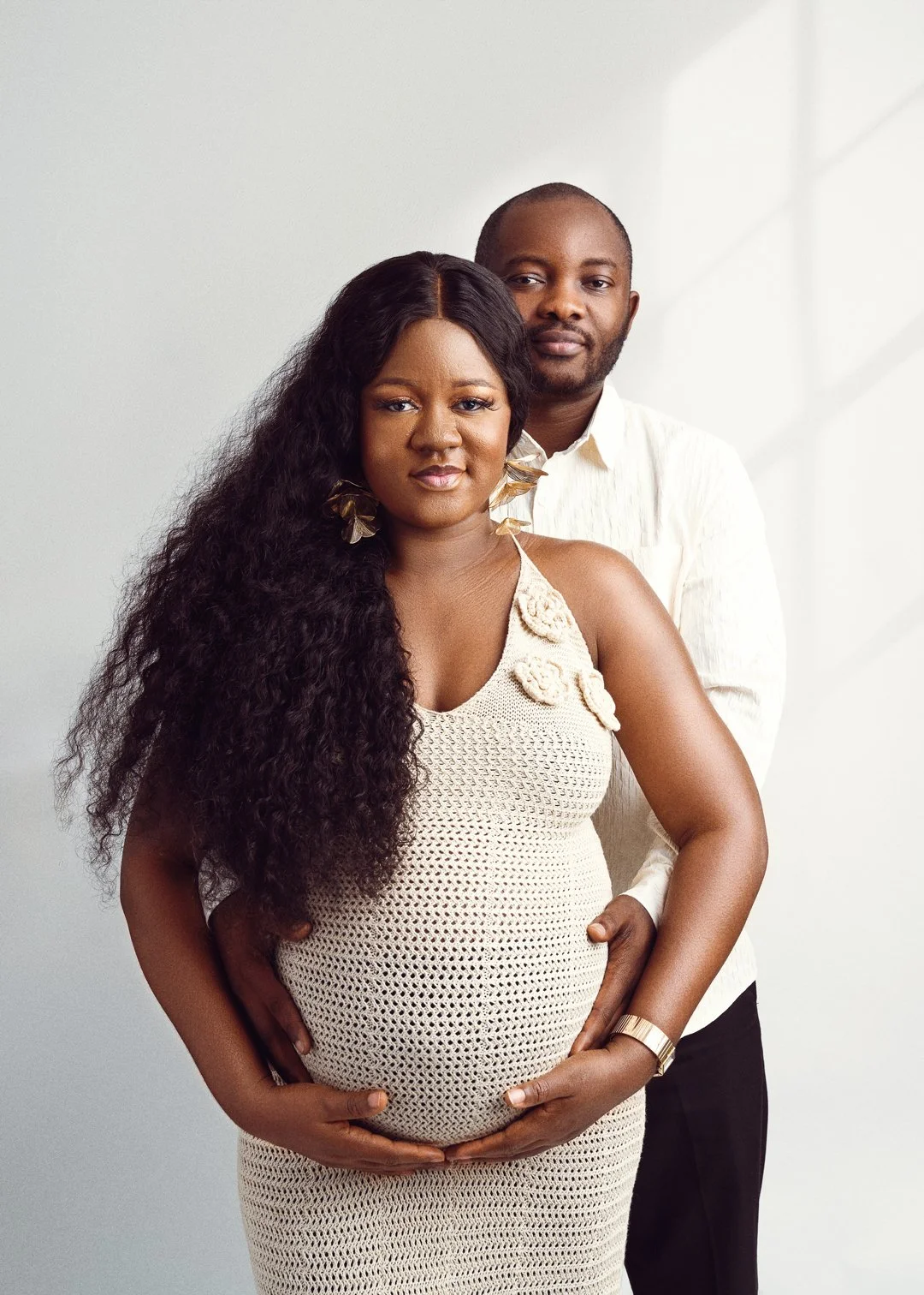 A pregnant woman with long curly hair and earrings standing in front, with a man standing behind her, both looking at the camera against a white background.