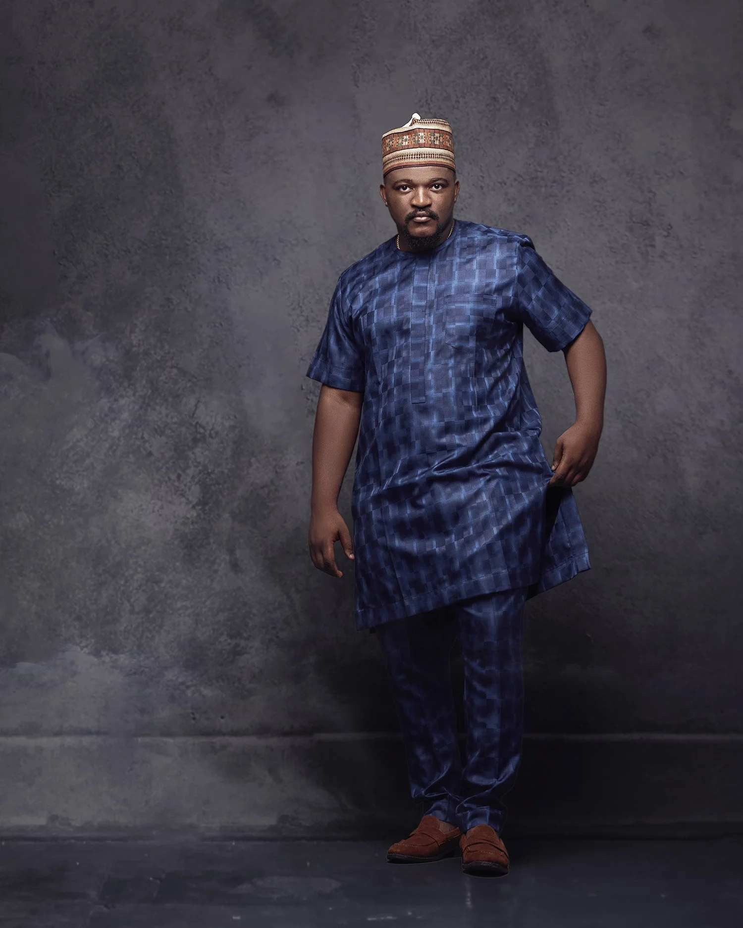 A man dressed in traditional Nigerian attire, consisting of a matching blue patterned agbada and trousers, with a traditional hat, posed against a textured gray background.