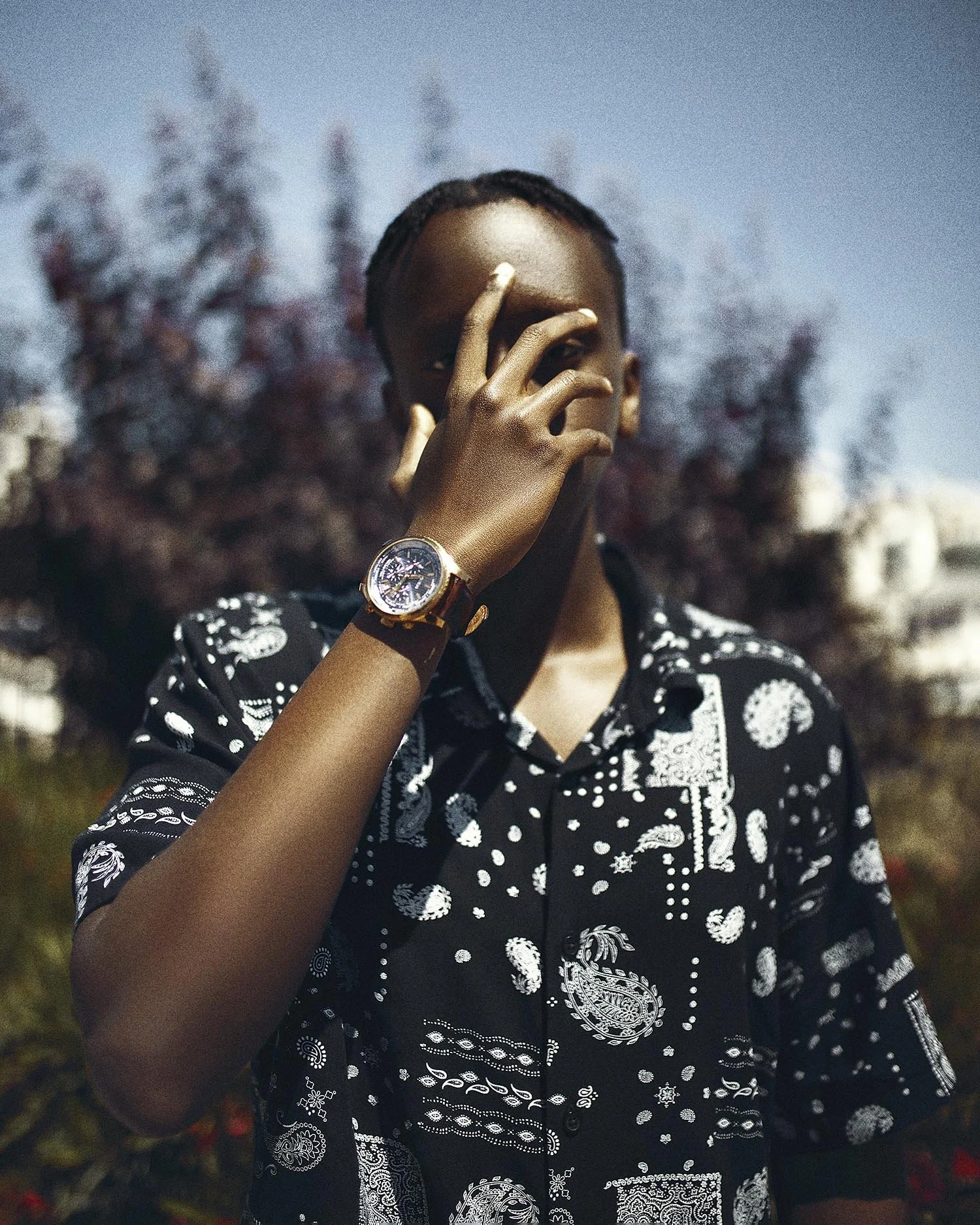 A young man in a black patterned shirt wearing a large wristwatch poses outdoors with the background of trees and buildings.