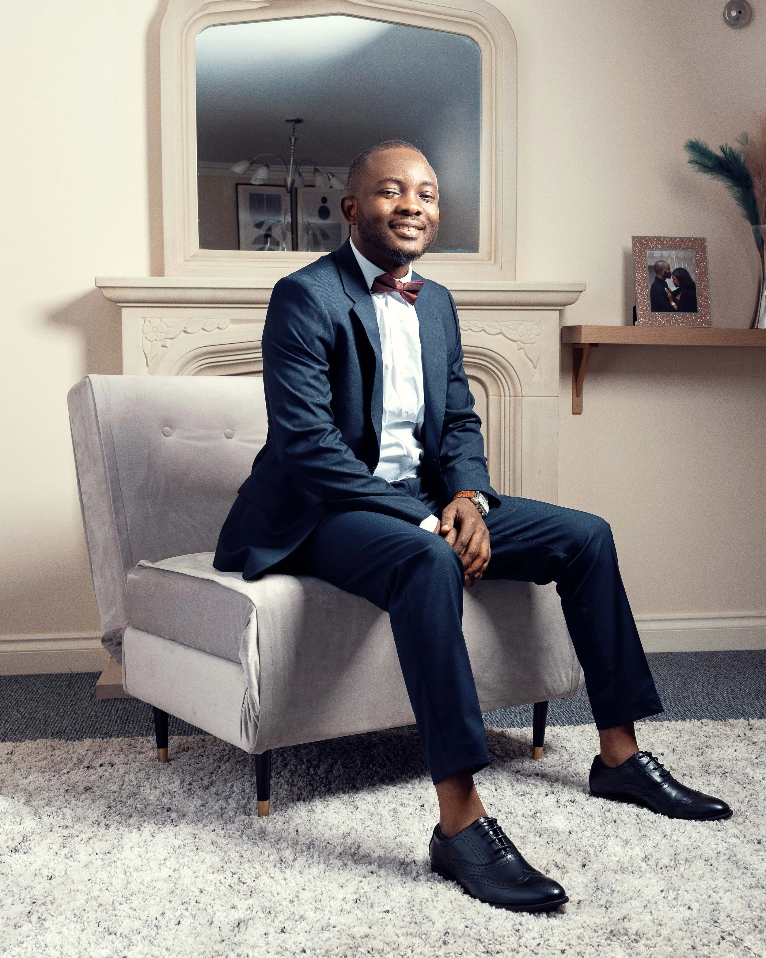 A young man in a navy blue suit, white shirt, and maroon bow tie, sitting on a light grey armchair in a living room, smiling at the camera. Behind him is a mirror over a fireplace and a shelf with a framed photo and plant.