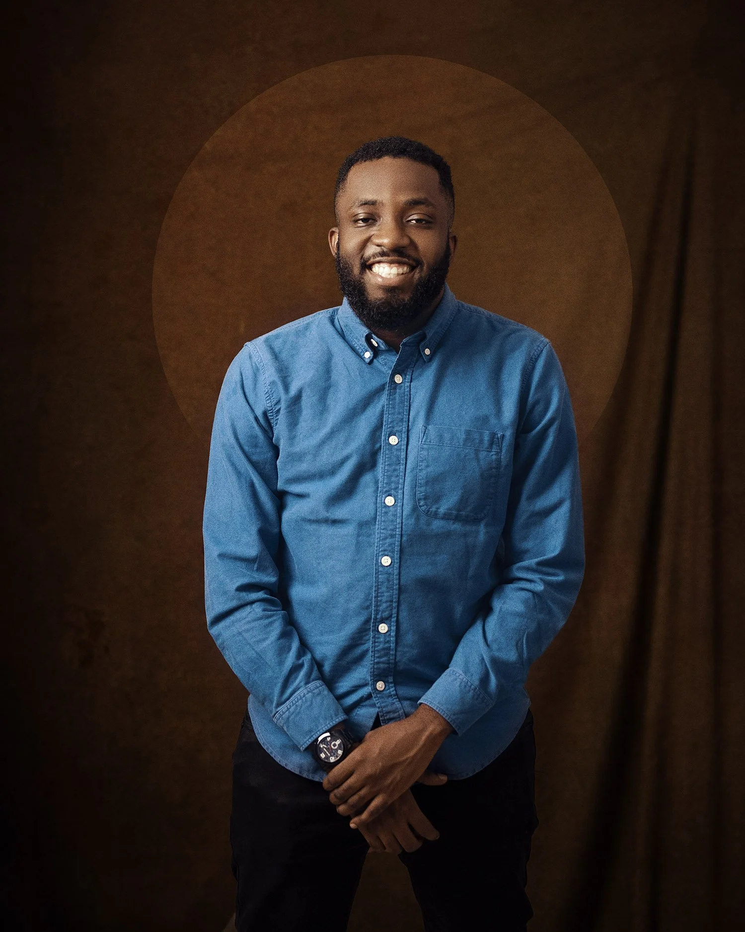 A smiling man with a beard wearing a blue button-up shirt and a watch, standing against a brown background.