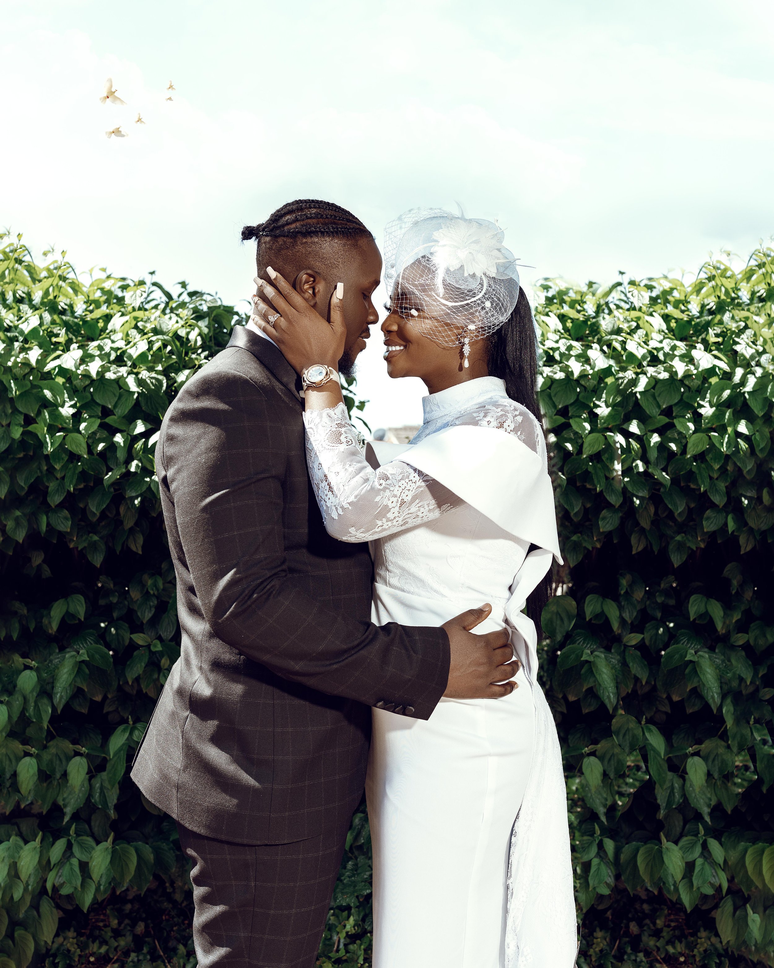 A couple dressed in wedding attire embracing outdoors, with green foliage and a partly cloudy sky in the background.