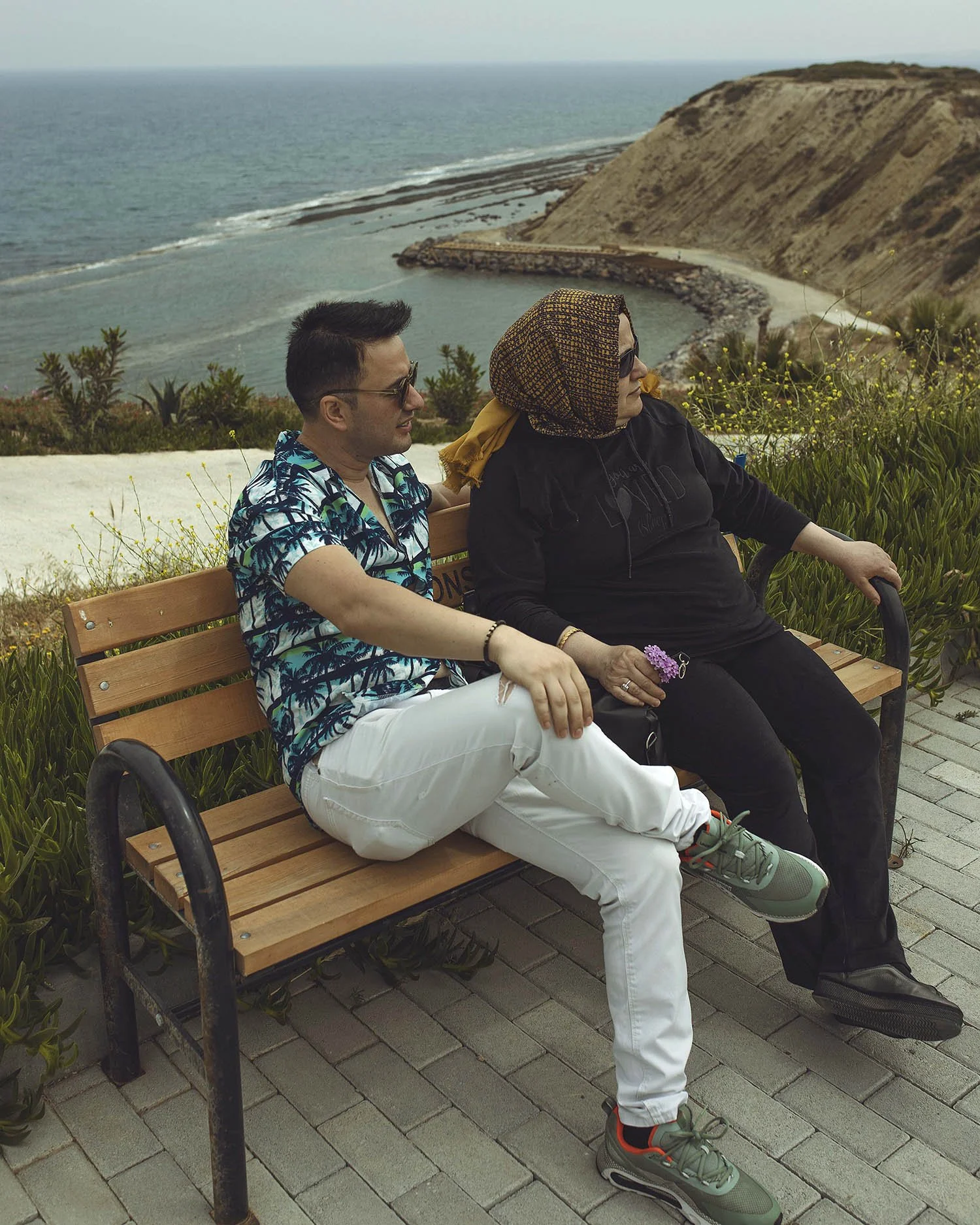 Two people sitting on a park bench overlooking the ocean and cliffs with greenery.