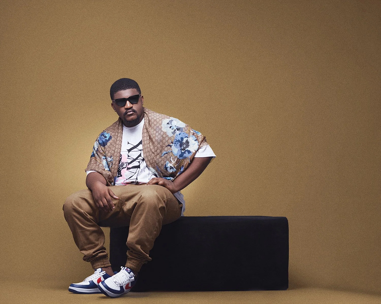 A man wearing sunglasses, a patterned jacket, a graphic t-shirt, tan pants, and sneakers, sitting on a black platform against a plain tan background.