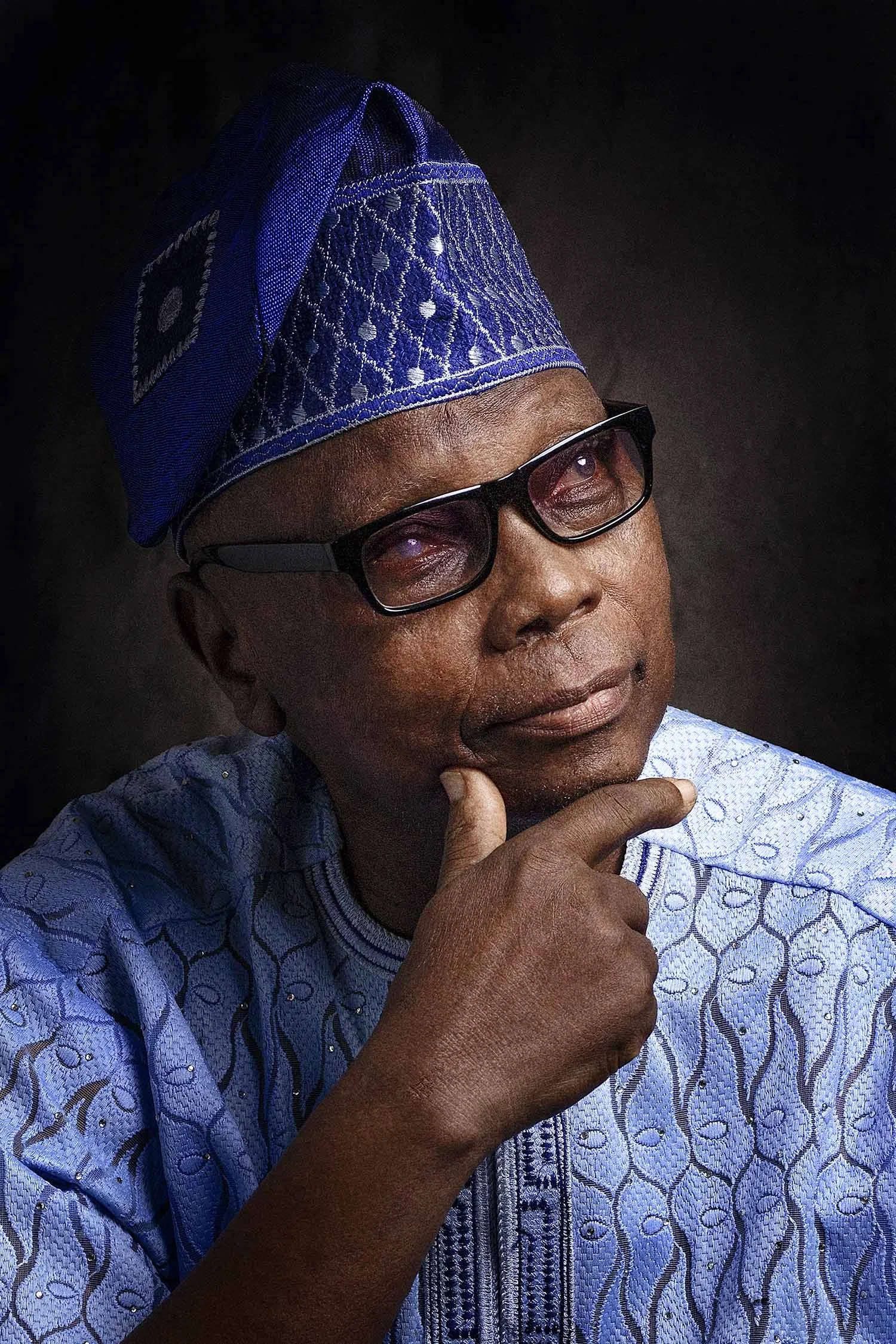 Portrait of a man wearing traditional Nigerian attire including a blue patterned agbada and a matching cap, with black glasses, against a dark background.
