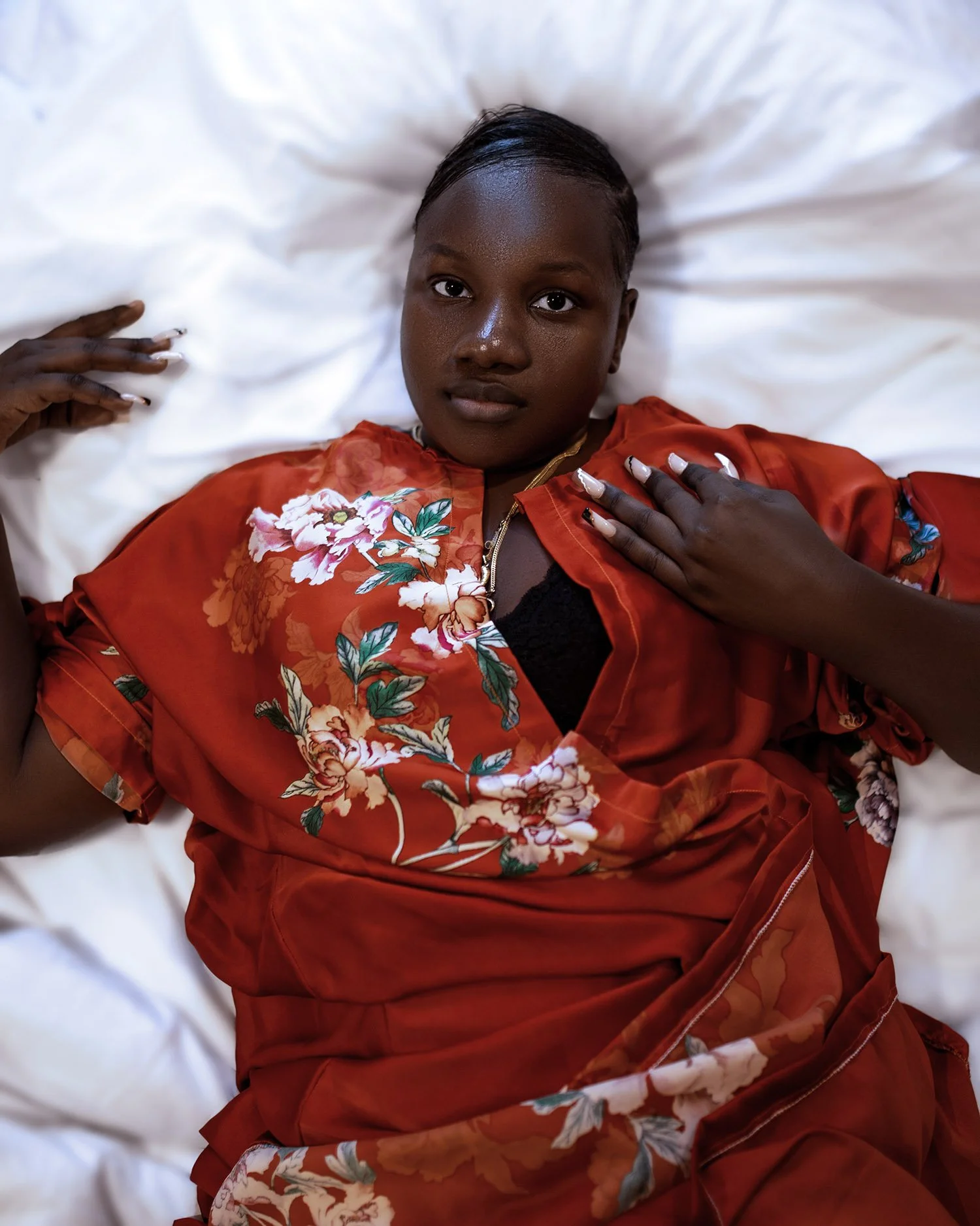 A woman with dark skin and short hair lying on a white pillow, wearing a red floral satin robe with green and pink flowers, looking at the camera.
