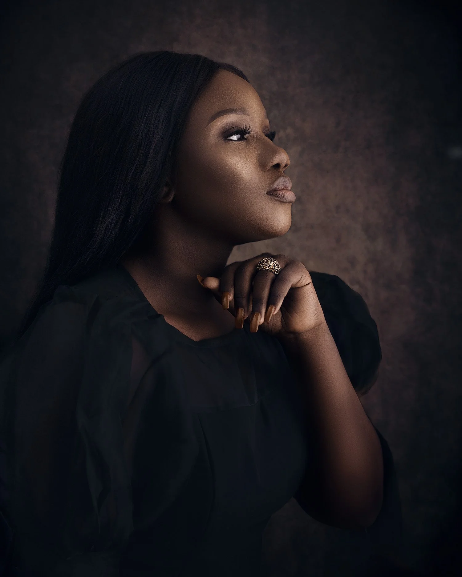 A portrait of a woman with dark hair, looking thoughtfully upward with her hand near her face, wearing a black top and a decorative ring.