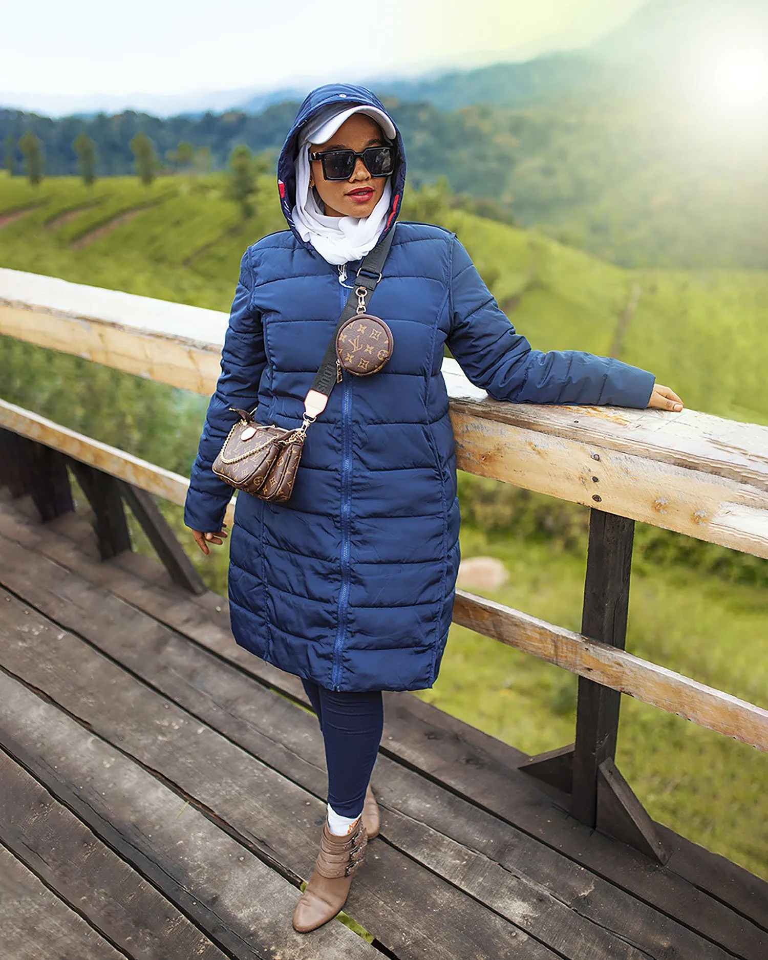A woman dressed in a blue puffy coat, dark sunglasses, and beige boots stands on a wooden bridge, looking at lush green rolling hills and trees in the background.