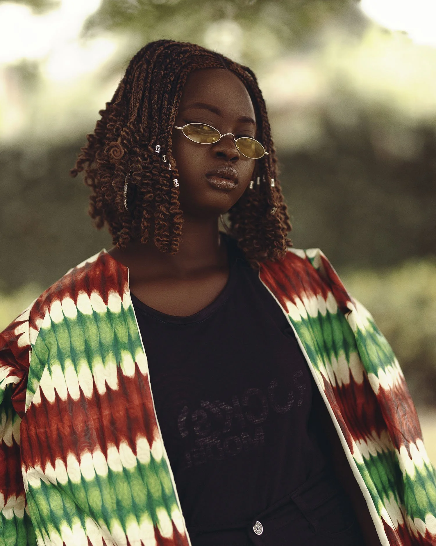 A woman with braided hair wearing yellow sunglasses, a black t-shirt, and a colorful patterned jacket outdoors.