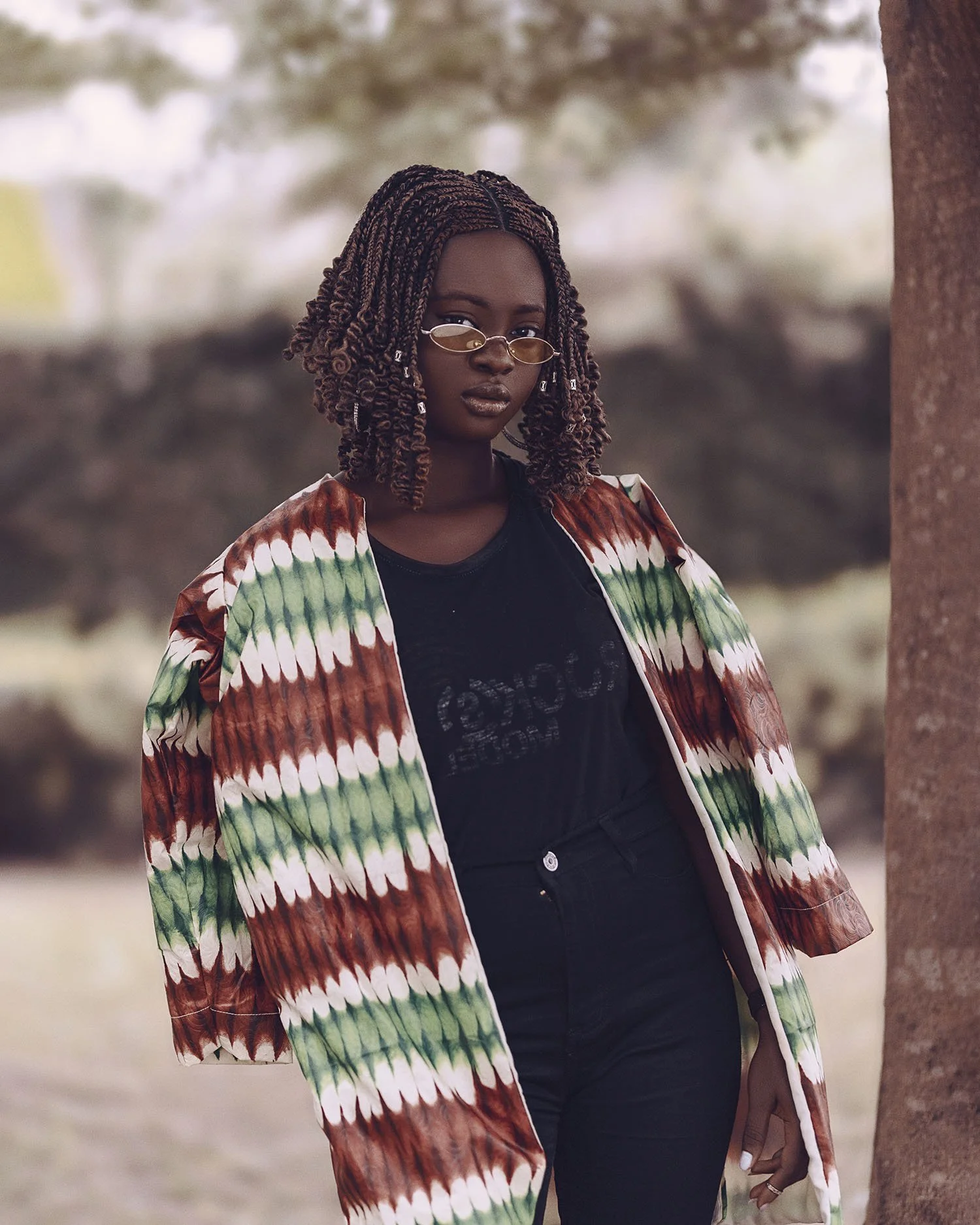 A woman with styled hair, wearing sunglasses, a colorful patterned jacket, and a black shirt, standing outdoors near a tree.