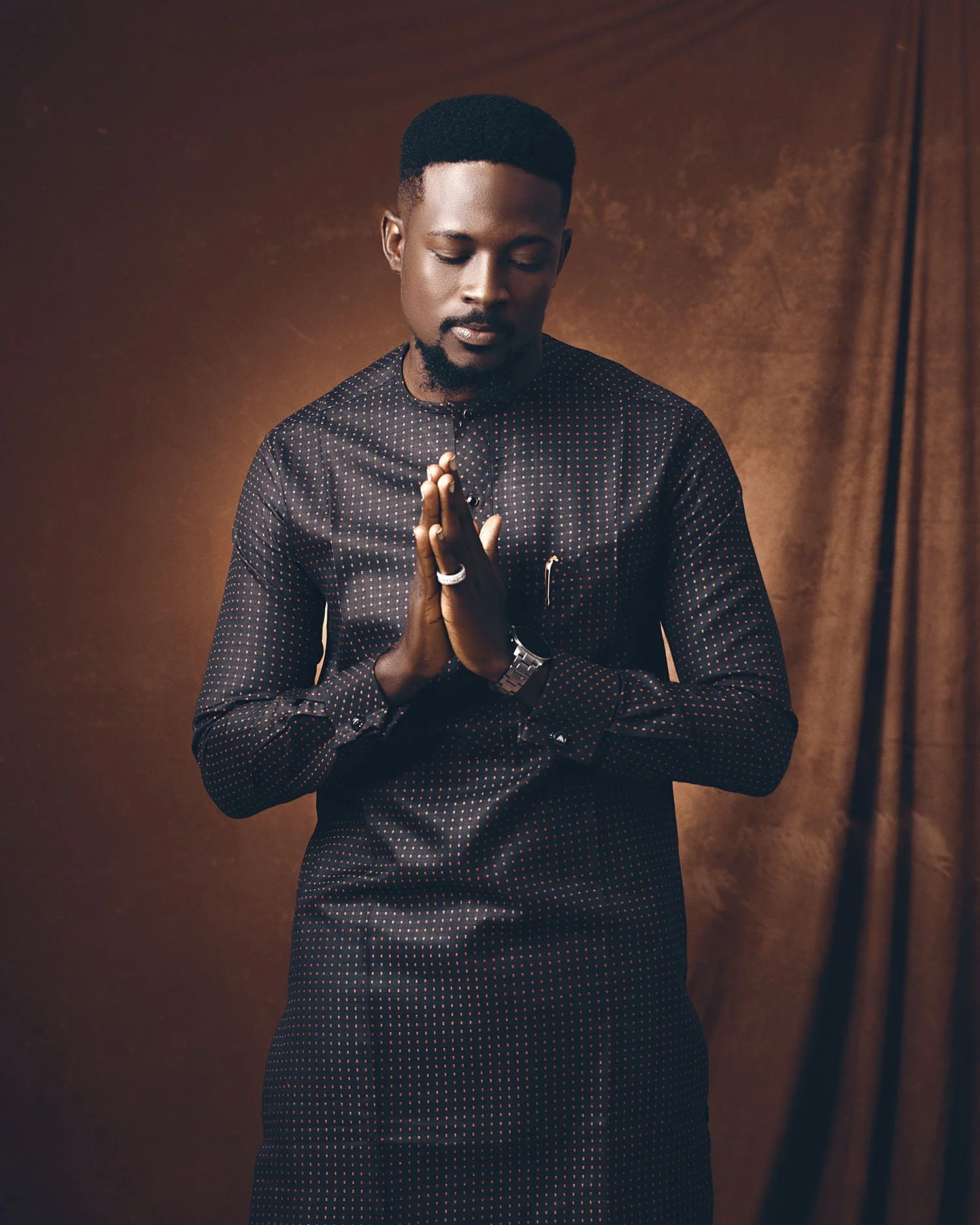 A young man with closed eyes, wearing a black shirt with small red and white polka dots, stands with hands pressed together in prayer or greeting against a brown background.