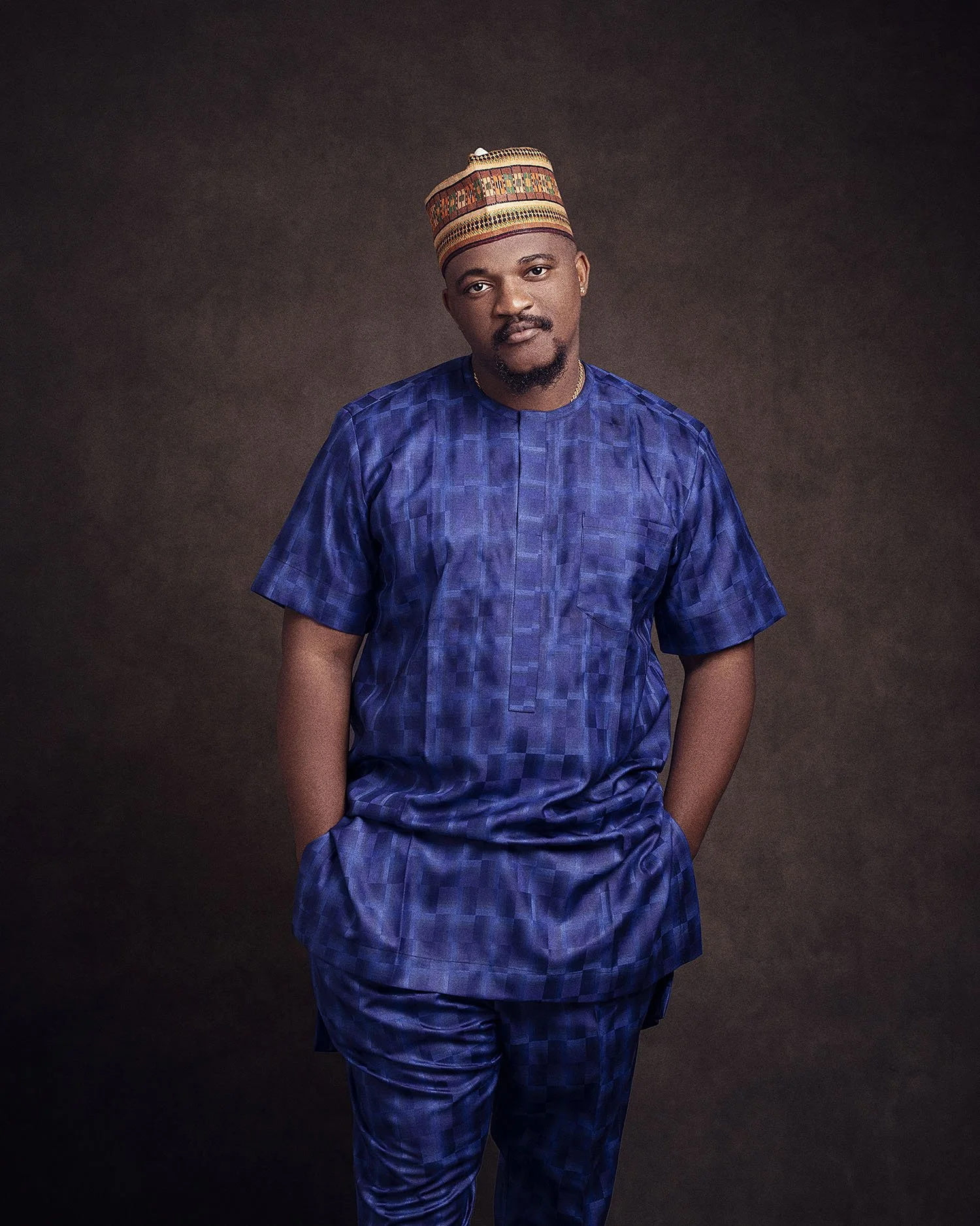 A man wearing traditional African attire in shades of blue, with hands in pockets, standing against a dark background.