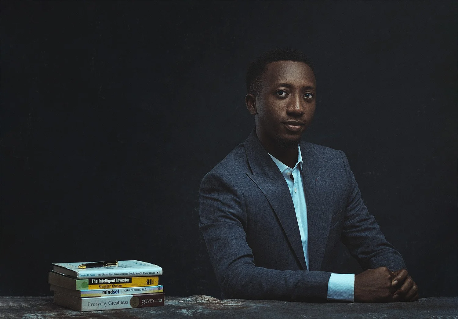 A young man in a blue suit sitting at a dark table with a stack of five books, black background.