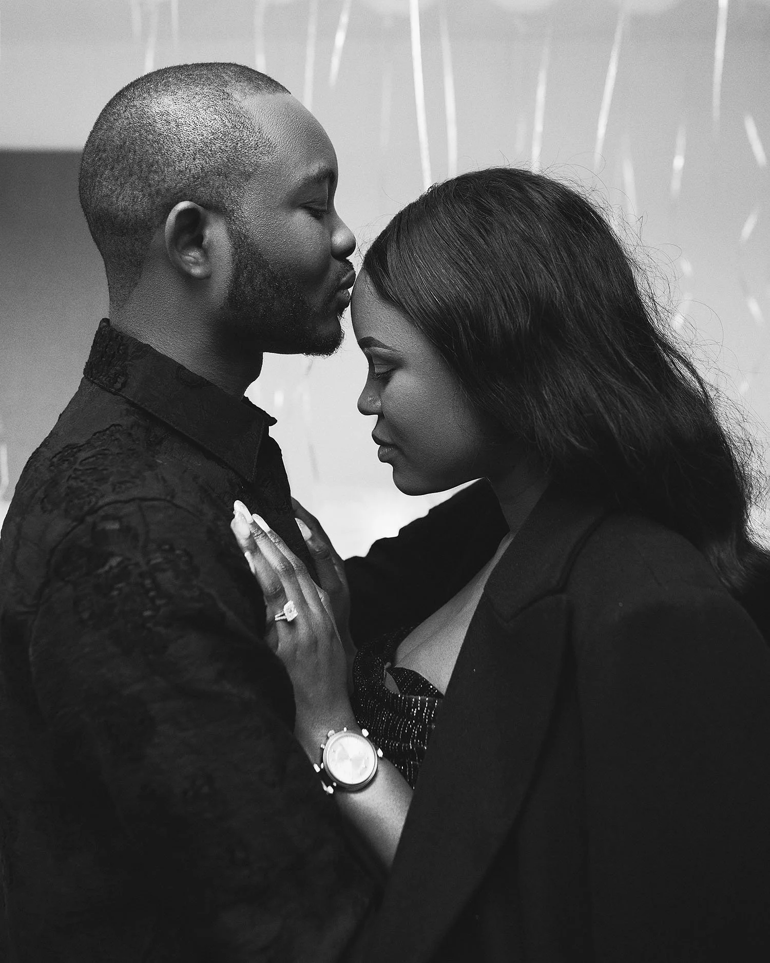 A black and white photo of a couple in an intimate pose, with their foreheads touching and eyes closed. The man is wearing a textured, lace-like jacket, and the woman is dressed in a blazer and a sparkly top. She is wearing a watch and an engagement 