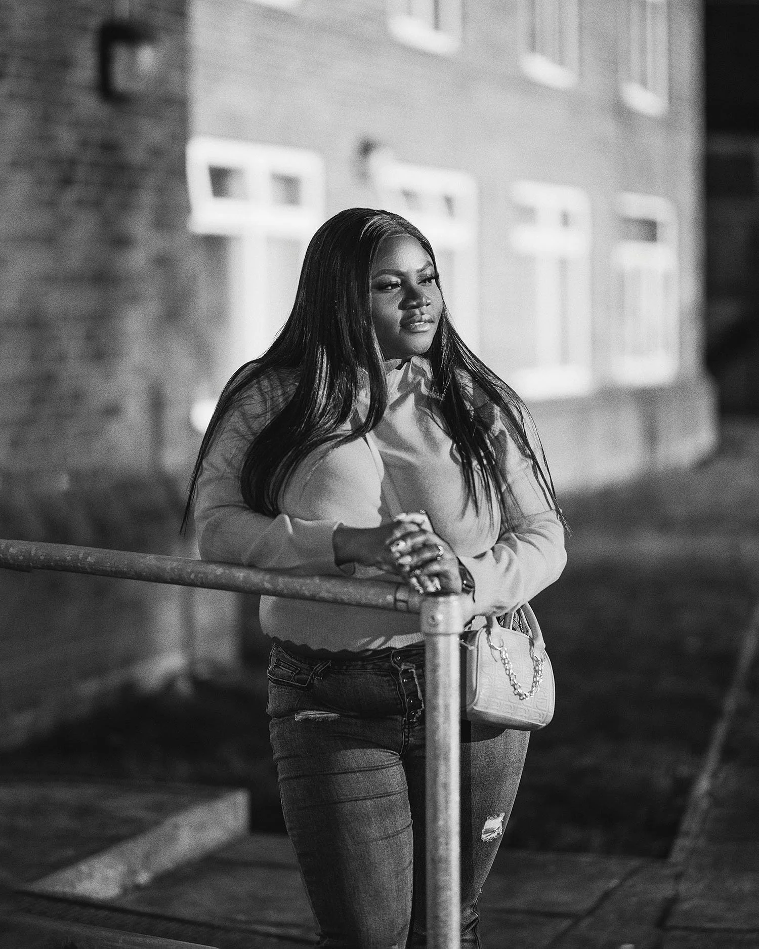 A woman with long dark hair and a light-colored top leaning on a metal railing outdoors at night, with a brick building in the background, captured in black and white.