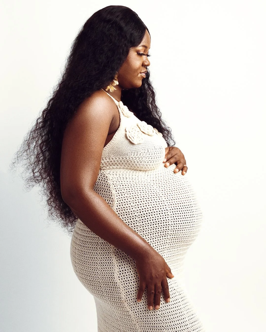 Pregnant woman wearing a beige crochet dress, standing sideways against a white background.
