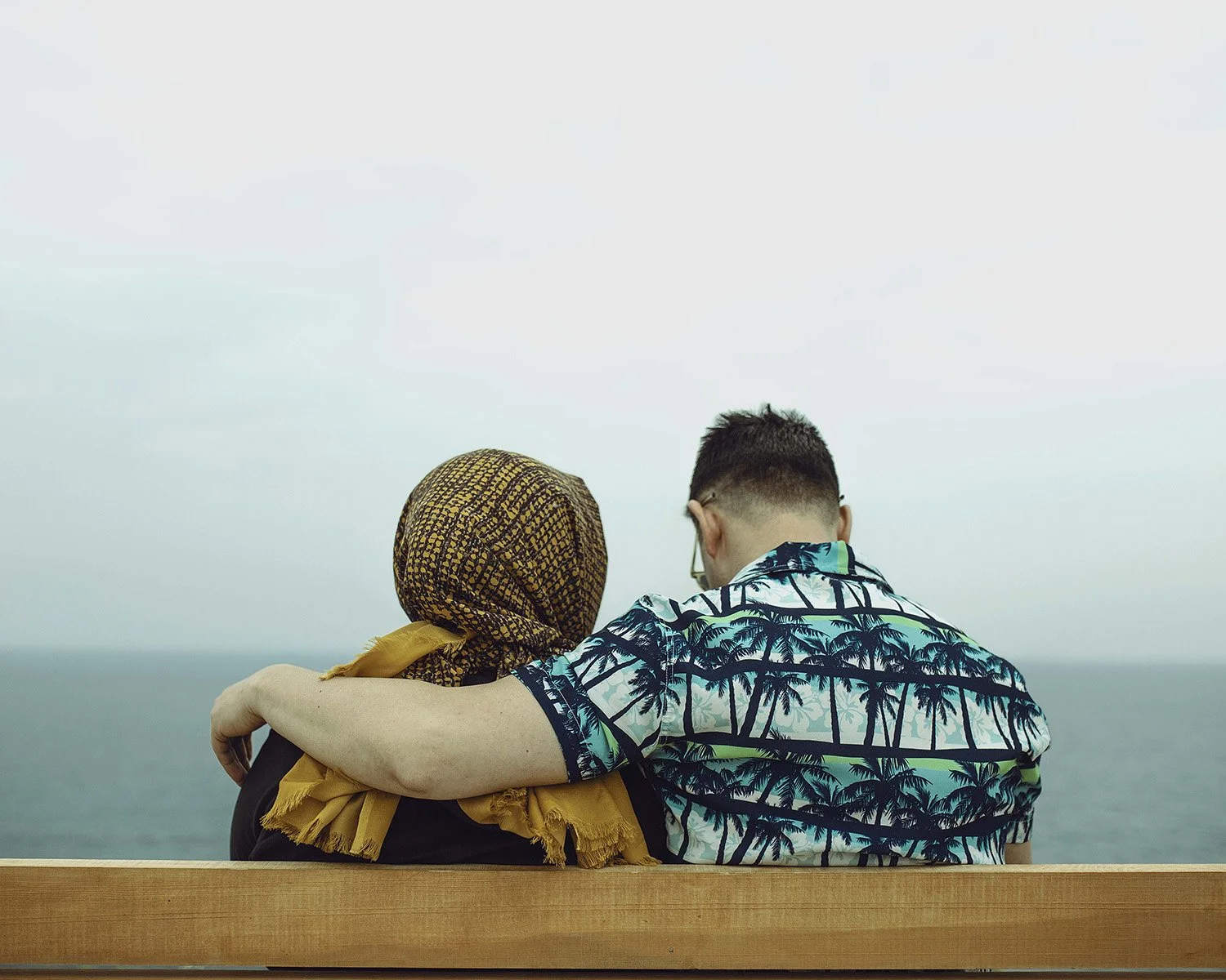 A couple sitting on a bench by the sea, with the man's arm around the woman's shoulder, viewed from behind, wearing casual summer clothing.