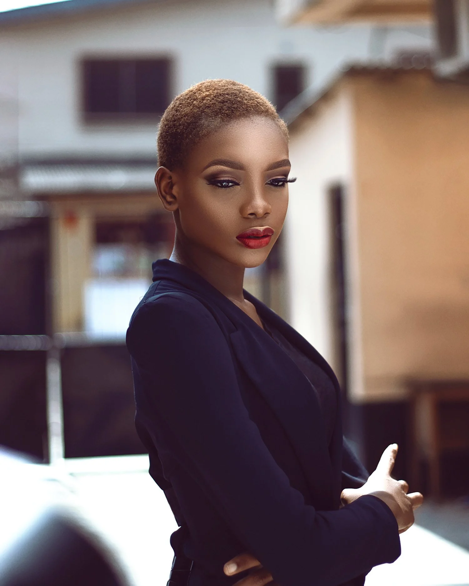 Portrait of a young woman with short, blonde hair, wearing a dark blazer, standing outdoors with blurred buildings in the background.