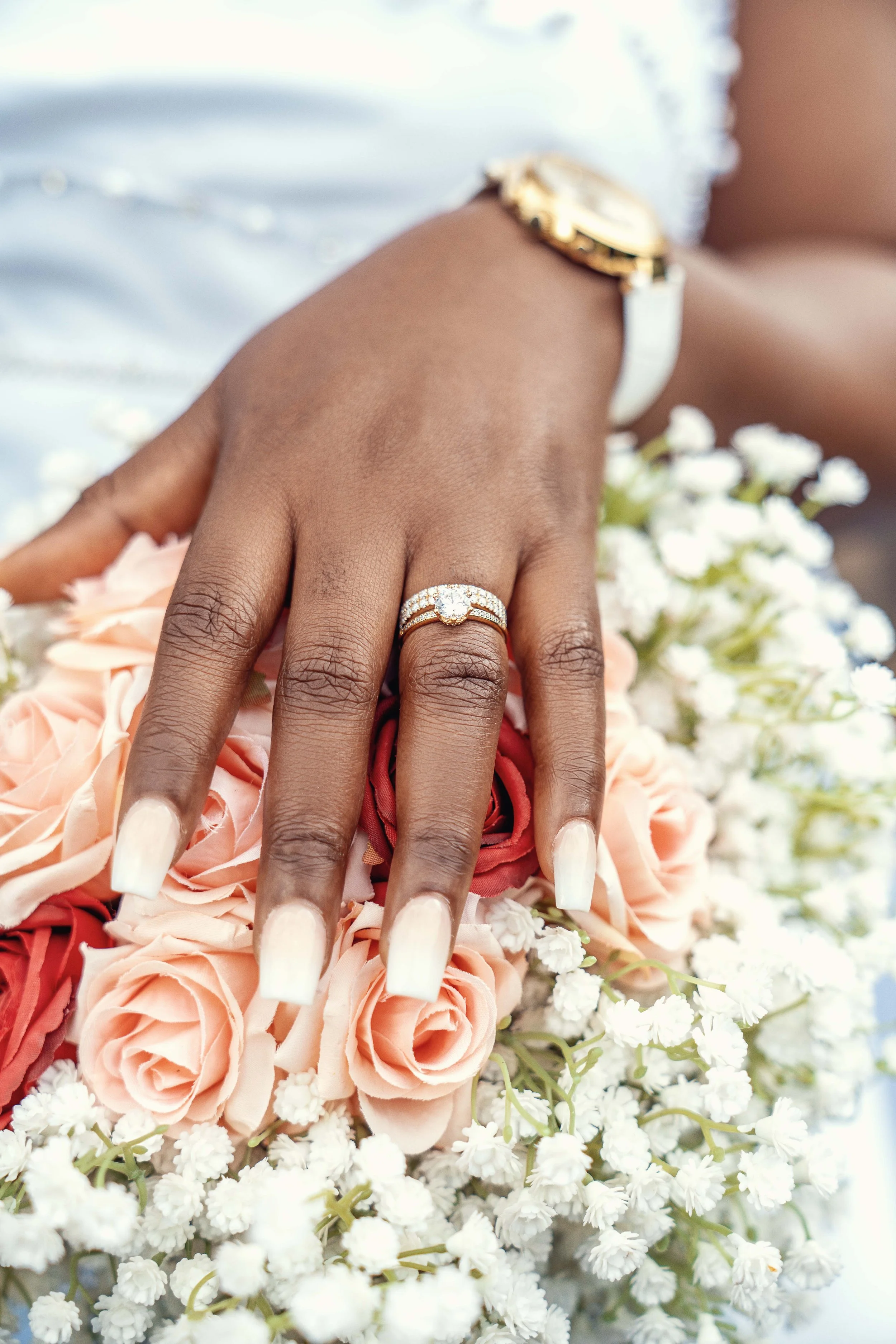 Close-up of a hand with a wedding ring resting on a bouquet of pink roses and white baby's breath flowers, with a blurred background.