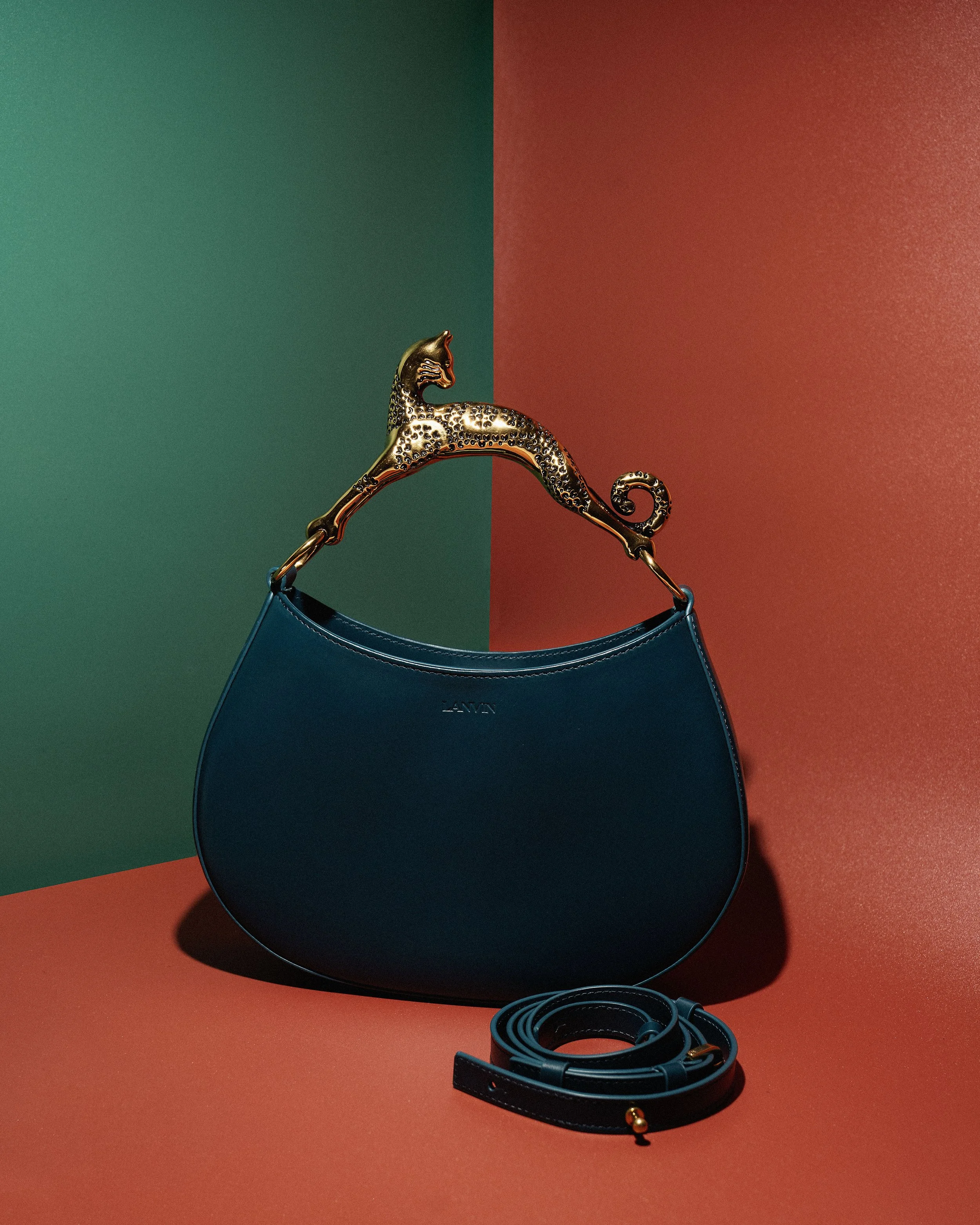 A blue handbag with a gold leopard-shaped handle and matching detachable strap, placed against a background with red and green sections.