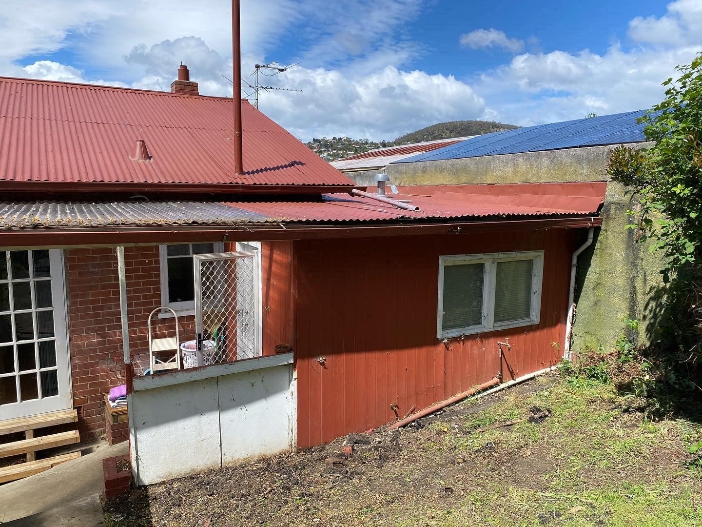Rear view of a house with a red metal roof, brick walls, a small balcony with white railing, and a window. There is a small patch of green grass and dirt, with some greenery on the right side and a hillside in the background under a partly cloudy sky