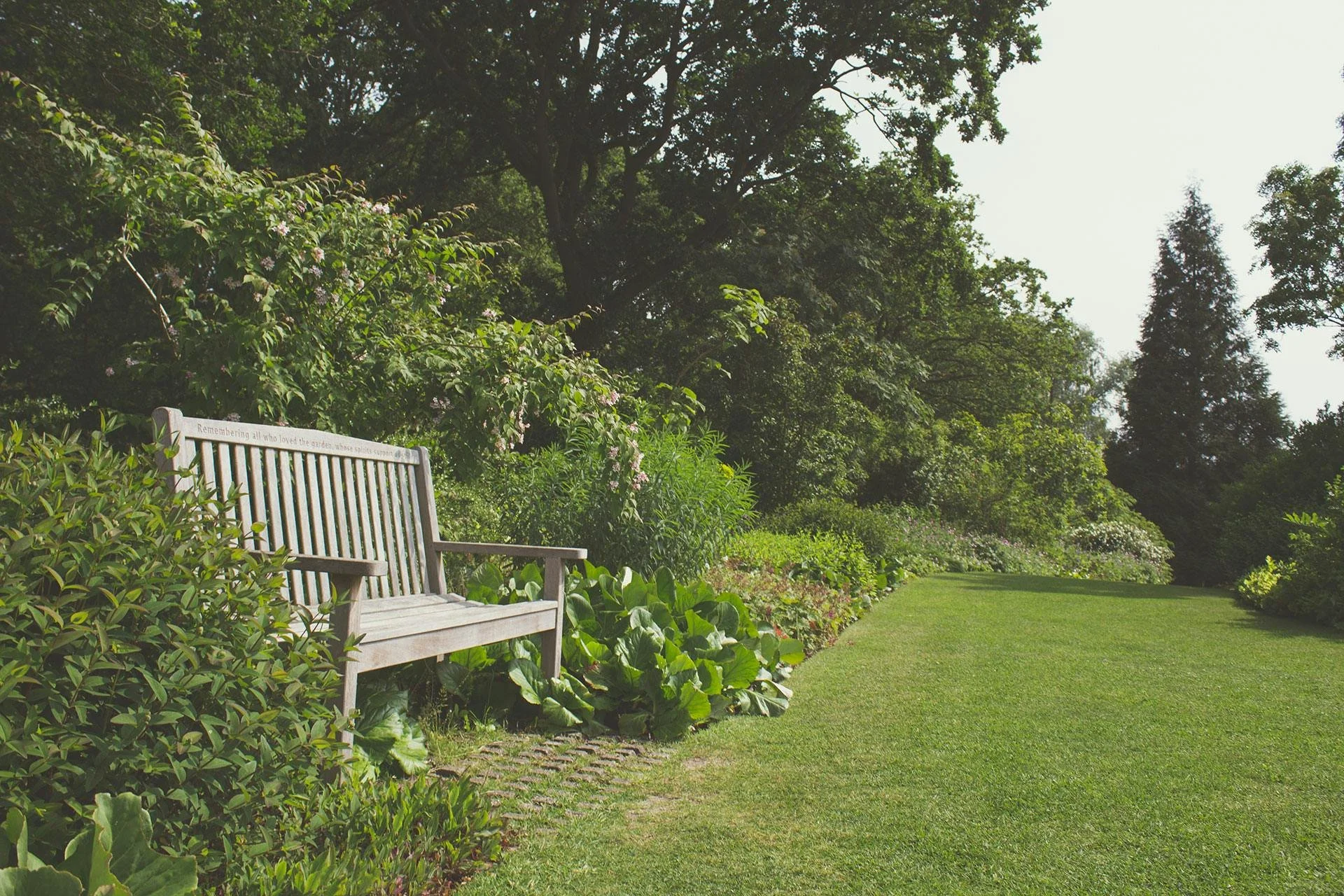 A wooden garden bench on a stone pathway next to a lush flower bed with green plants and colorful flowers, bordered by a well-maintained grassy lawn, with tall trees and dense foliage in the background on a sunny day.