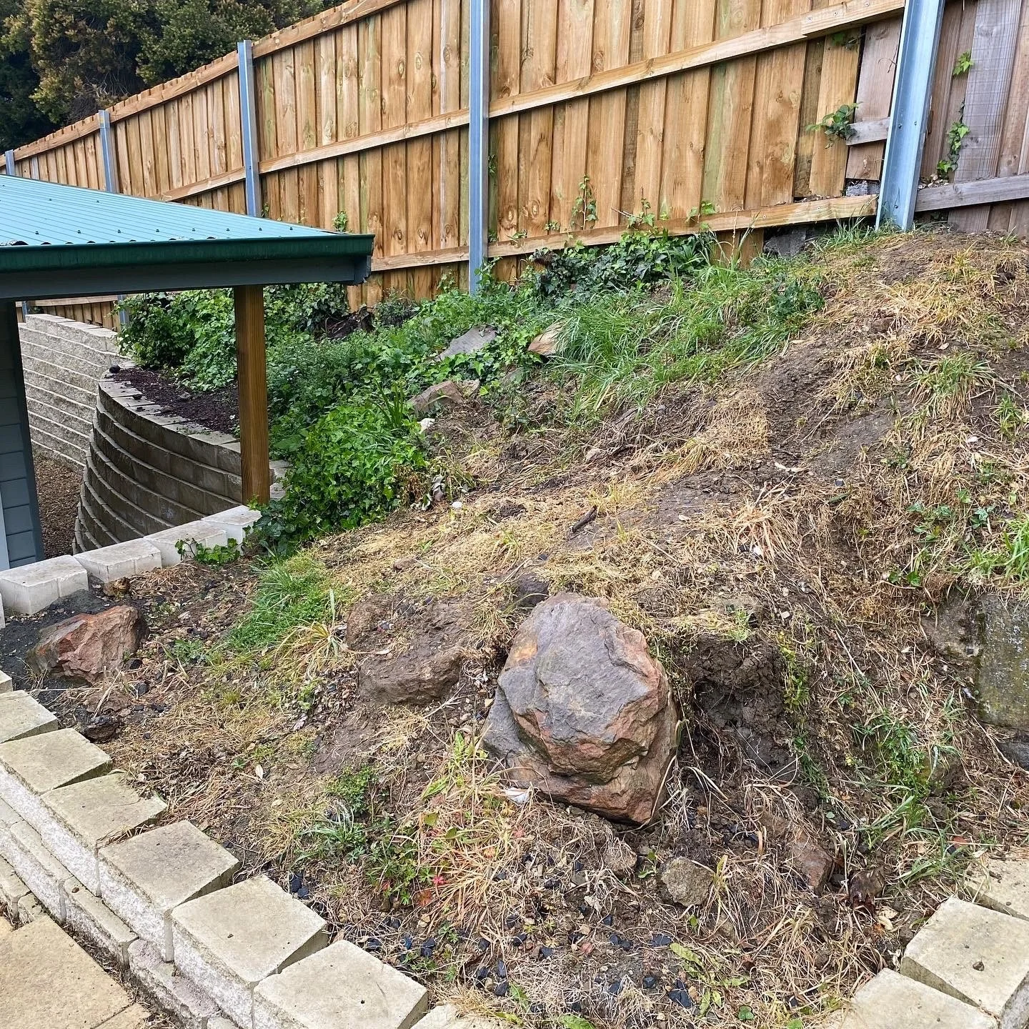 A sloped backyard area with a wooden fence, some plants, and a retaining wall made of bricks.