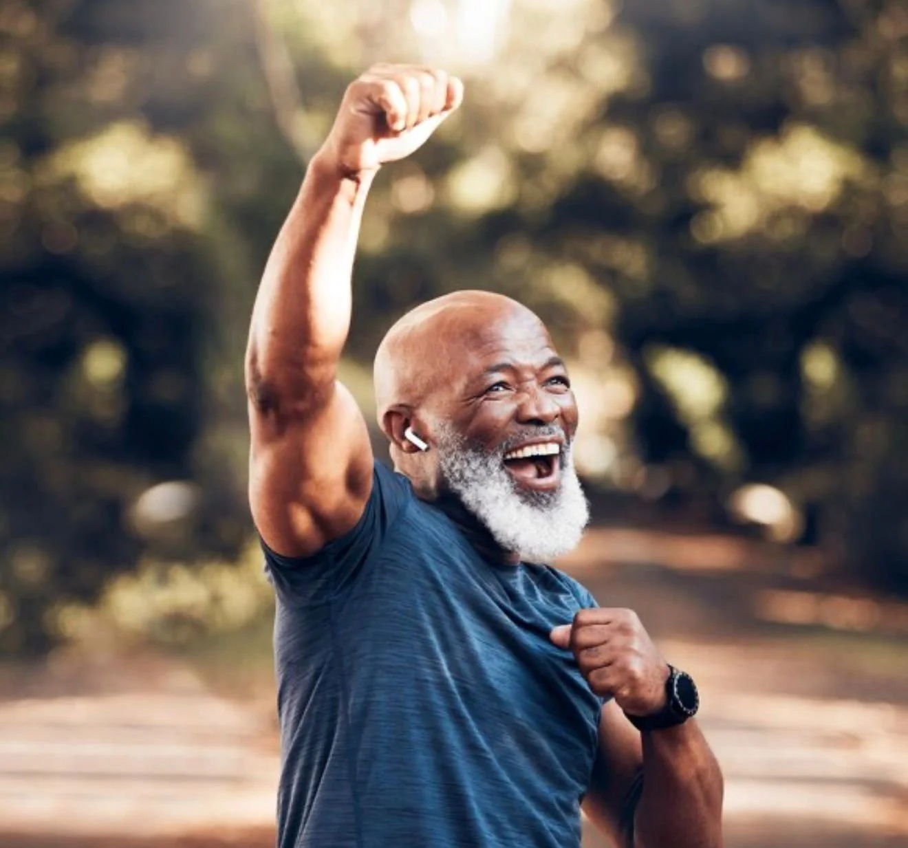 An elderly man with a gray beard and bald head smiling with enthusiasm, raising a fist in a gesture of victory or celebration, outdoors with a blurred natural background.