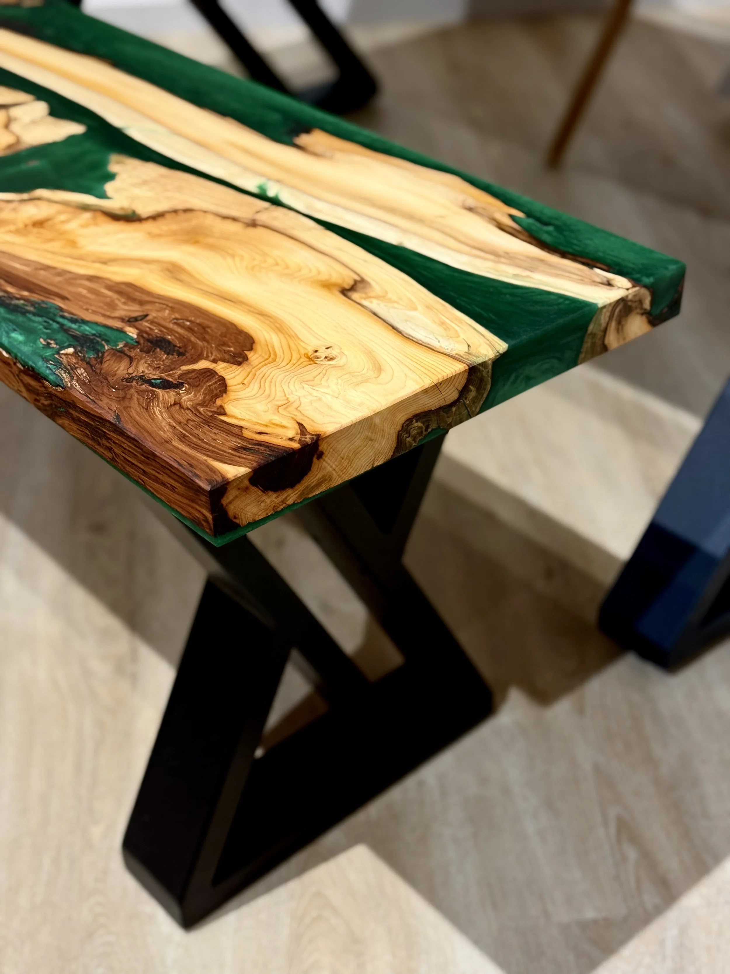Close-up of a wooden table with a combination of natural wood grain and green resin inlays, supported by black metal legs.