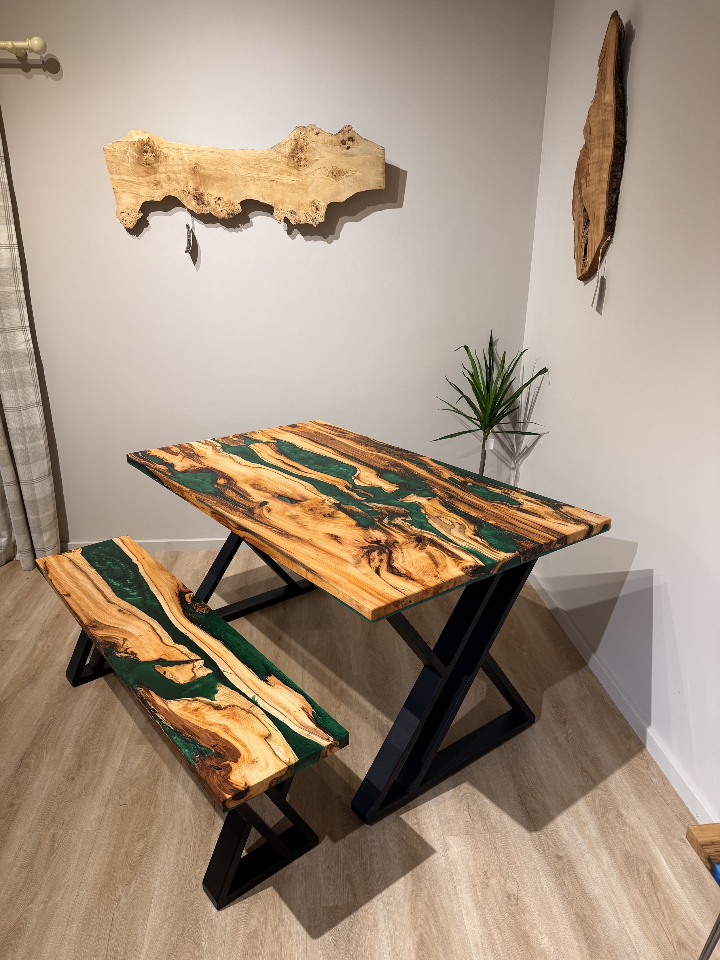 A wooden table with a matching bench and wall art, all featuring a natural wood and green resin design, in a room with light-colored wood flooring and a potted plant.