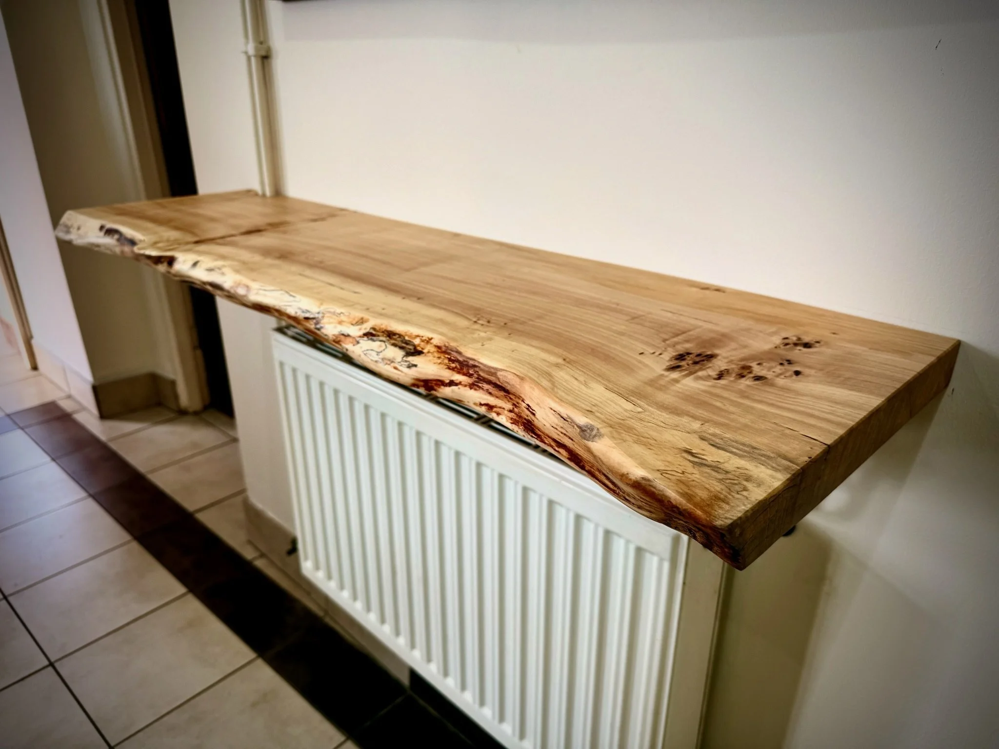 Wooden countertop with natural edge design, placed above a white radiator cover in a room with tiled floor.